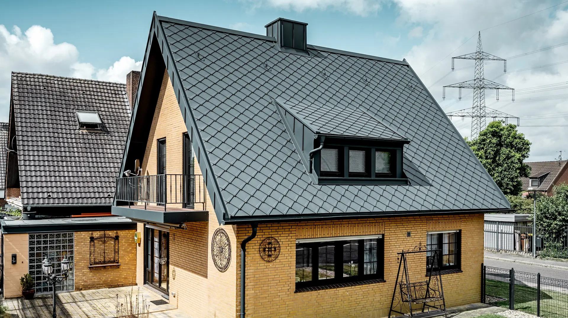 Rodinný dom zo žltého klinkeru po renovácii strechy s PREFA strešnou šablónou 29 × 29 v tmavošedé farbe; novo pokrytá strmá sedlová strecha s kosoštvorcovým vzorom, integrovanou vikierovou strechou a komínom, terasa a záhrada v popredí.