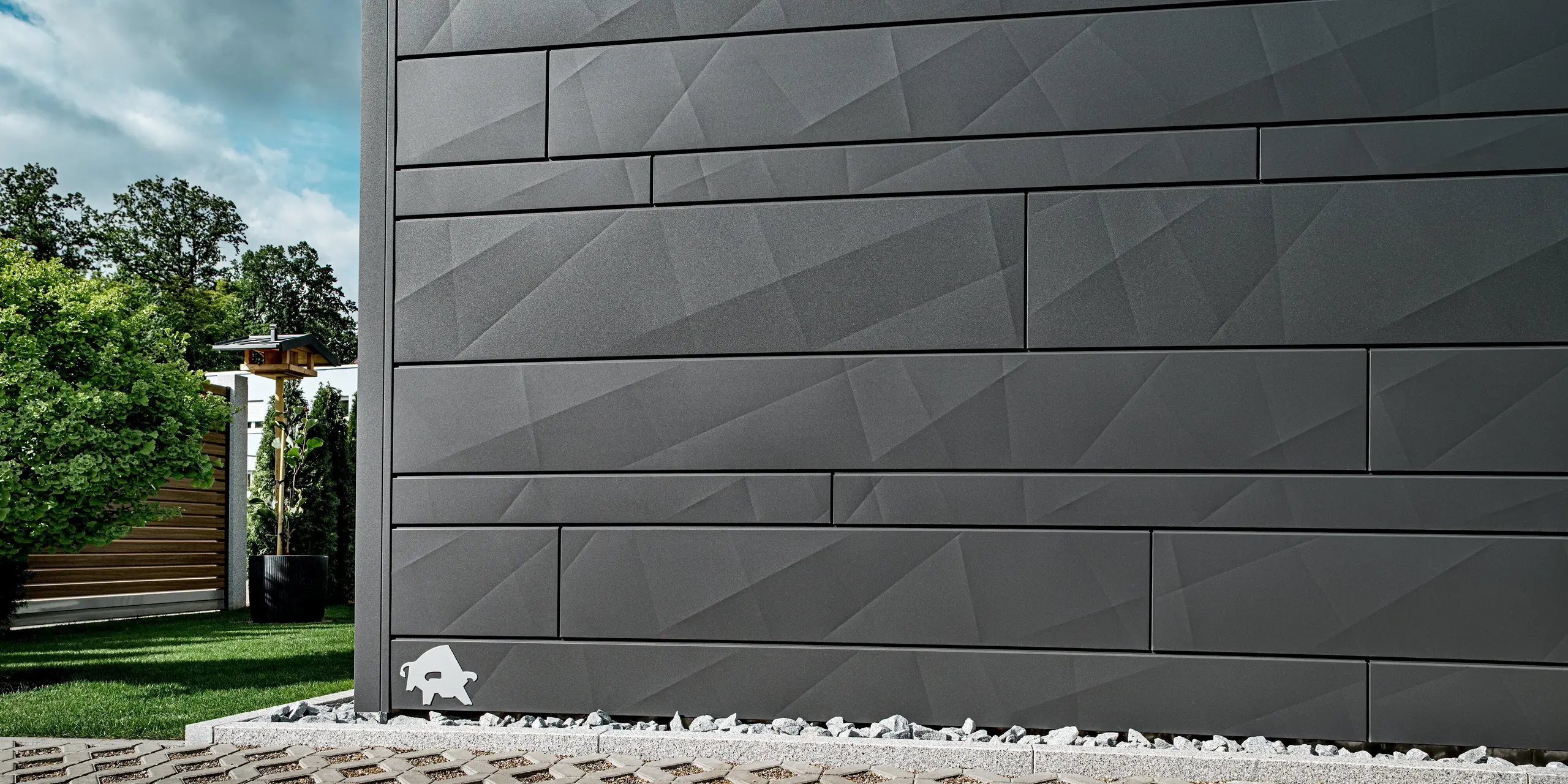 Detailansicht der PREFA Siding.X Aluminiumfassade in der Farbe P.10 Dunkelgrau an einem modernen Einfamilienhaus in Heidenheim. Die klar strukturierten Paneele mit variierenden Breiten und markanten Kantungen erzeugen ein prägnantes, geometrisches Muster, das je nach Lichteinfall spannende Schattenspiele bildet. Im Vordergrund sind helle Ziersteine und ein gepflegter Gartenbereich zu sehen, im Hintergrund Bäume und eine Holzstruktur. Die langlebige und witterungsbeständige Aluminiumoberfläche unterstreicht die hochwertige und moderne Fassadengestaltung. Am linken Rand der Fassade ist das bekannte PREFA-Logo, der Stier, zu erkennen.