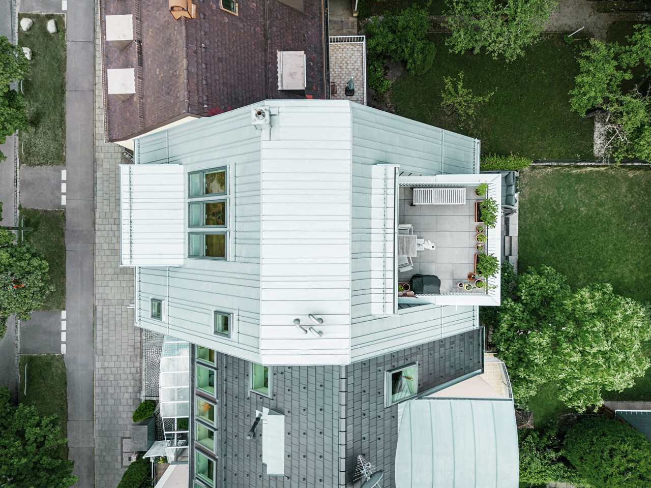 Letecký pohľad na rohový radový dom s modernou rekonštrukciou. Dom má strechu a fasádu horného poschodia zo striebornej metalízy Prefalz hliníka, ktorá zvýrazňuje horizontálne línie. Strešná terasa s vonkajším nábytkom a rastlinami vytvára mestskú zelenú oázu. Fasáda suterénu je v tmavšom tóne, čo vytvára príťažlivý kontrast. Dom je obklopený udržiavanou zeleňou a spevnenými chodníkmi, ktoré podčiarkujú jasnú štruktúru stavby.