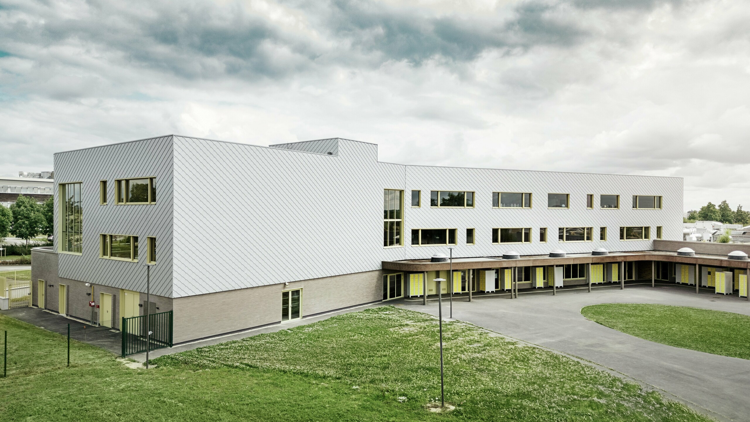 Na obrázku je zadná časť školy Collège Léontine Dolivet. V popredí sa nachádza veľké asfaltové školské ihrisko s jasnými líniami a otvorenými plochami, ktoré sa dajú využiť na prestávky a voľnočasové aktivity. Niekoľko zelených plôch oživuje okolie. V pozadí sa týči moderná budova školy s výraznou hliníkovou fasádou z fasádnych šablón, ktoré odrážajú svetlo a vytvárajú dynamický efekt. Niekoľko veľkých okien zaisťuje svetlosť v interiéri. Scéna je nasnímaná za denného svetla, pri mierne zamračenom nebi, čo zvlášť zdôrazňuje kovový povrch.