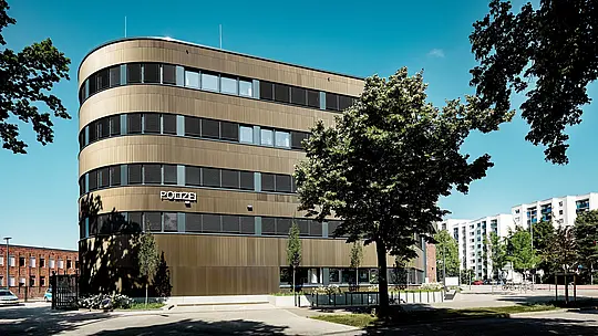 Dynamický architektonický záber policajnej stanice č. 43 v Hamburgu: Zakrivená nová budova s hliníkovou fasádou PREFA z extrudovaných profilov „Zacke“ v odtieni strednej bronzy priťahuje ako výrazný prvok v mestskom prostredí pozornosť všetkých okoloidúcich. Jemne štruktúrovaný povrch vytvára živé efekty svetla a tieňa medzi horizontálnymi pásmi okien a zdôrazňuje plynulé línie fasády. Hliník PREFA presviedča vysokou odolnosťou proti korózii, dlhou životnosťou a nízkymi nárokmi na údržbu – je ideálny pre udržateľné, funkčné a architektonicky precízne riešenia fasád.