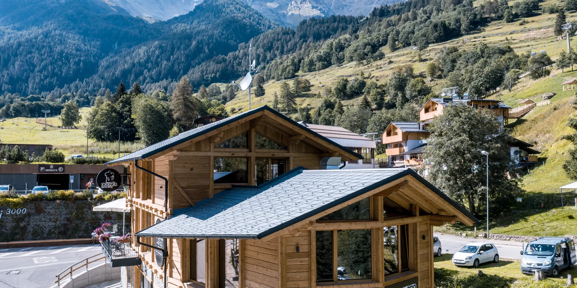 Detailný záber na pôvabný drevený apres-ski bar v alpskej krajine, pokrytý hliníkovými strešnými šindľami PREFA DS.19 vo farbe P.10 kamenná šedá. Malebná stavba má terasu, na ktorej si hostia môžu vychutnávať úchvatný výhľad na talianske hory, pričom miesto dokonale zapadá do okolitej architektúry a sviežich horských lúk.