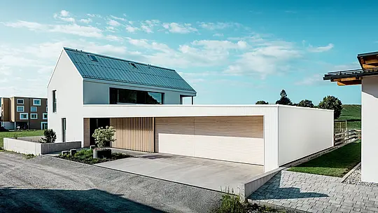 Na obrázku je moderný rodinný dom vo Freistadte, ktorý sa vyznačuje jednoduchými geometrickými tvarmi a kombináciou čisto bielej hliníkovej fasády PREFALZ a drevených obkladov. Drevený obklad na garážových dverách vytvára teplý, prirodzený kontrast a dokonale zapadá do moderného dizajnu. Strecha, tiež pokrytá PREFALZ, zaisťuje hladký povrch odolný voči poveternostným vplyvom. Táto kombinácia ľahko udržiavateľného hliníka a dreva vytvára esteticky príťažlivé a funkčné riešenie pre udržateľnú architektúru. Rozľahlý zelený exteriér a jasná obloha v pozadí podčiarkujú modernú kvalitu života, ktorú tento dom ponúka.