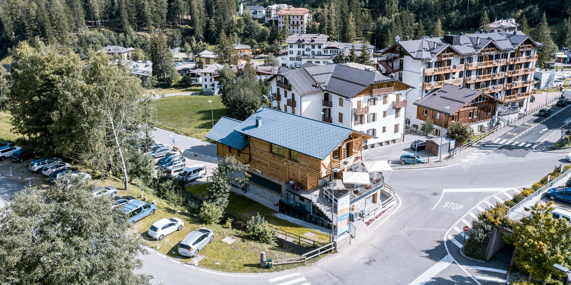 Pohľad z vtáčej perspektívy na živú križovatku v alpskej dedinke s nápadným après-ski barom so strešnými šindľami PREFA DS.19 vo farbe P.10 kamenná šedá. Kombinácia tradičnej drevenej konštrukcie a moderných hliníkových strešných prvkov, obklopená kulisou husto zalesnených kopcov a zmesou moderných a tradičných budov, je dôkazom úspešnej symbiózy inovácie a tradície v srdci talianskych Álp.