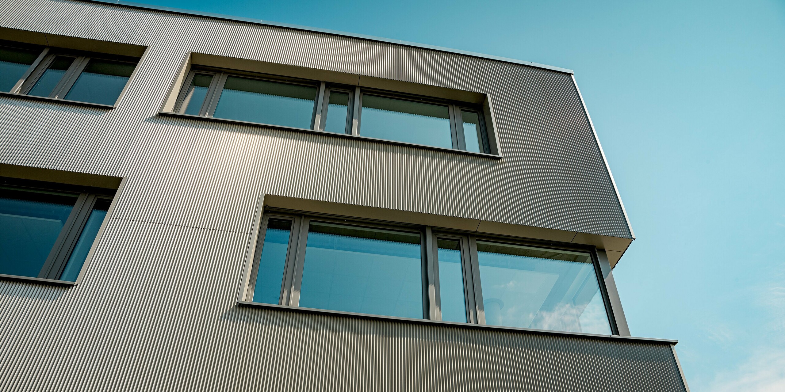 Das Bild zeigt eine Detailaufnahme der Aluminiumfassade des Festo-Gebäudes in Langenhagen, die mit PREFA Strangpressprofilen in Zackenform ausgestattet ist. Die vertikale Struktur der Profile verleiht der Fassade eine markante, texturierte Optik, die das Gebäude sowohl modern als auch dynamisch erscheinen lässt. Besonders beeindruckend ist die klare Linienführung und die präzise Integration der Fenster, die eine harmonische Balance zwischen Architektur und Funktionalität bieten. Der Einsatz der robusten und langlebigen PREFA Produkte sorgt für eine ästhetische und witterungsbeständige Fassade, die den hohen Anforderungen an Design und Nachhaltigkeit gerecht wird.