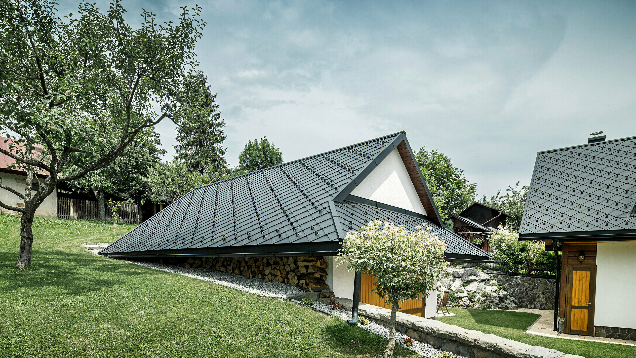 Prístavba tradičného rodinného domu v Starej Bystrici, Slovensko, so strechou z PREFA strešných šablón 29 × 29 vo farbe P.10 čierna. Robustná hliníková strecha s pultovým vzhľadom chráni budovu a harmonicky zapadá do zeleného vidieckeho prostredia. V popredí je vidieť udržiavaný trávnik a strom, čo dodáva celkovému obrazu idylickú a prívetivú atmosféru.