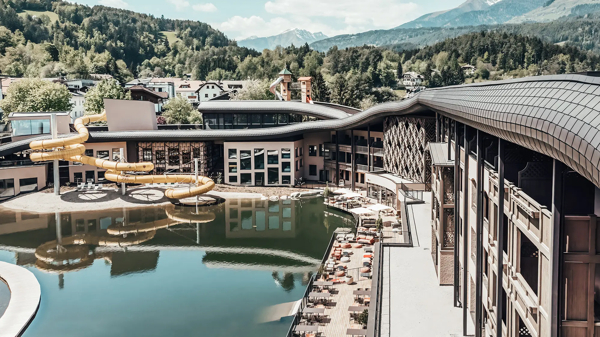 Überblick auf das Hotel mit See und Wasserrutsche. Im Hintergrund ist hügelige und bewaldete Fläche zu sehen.