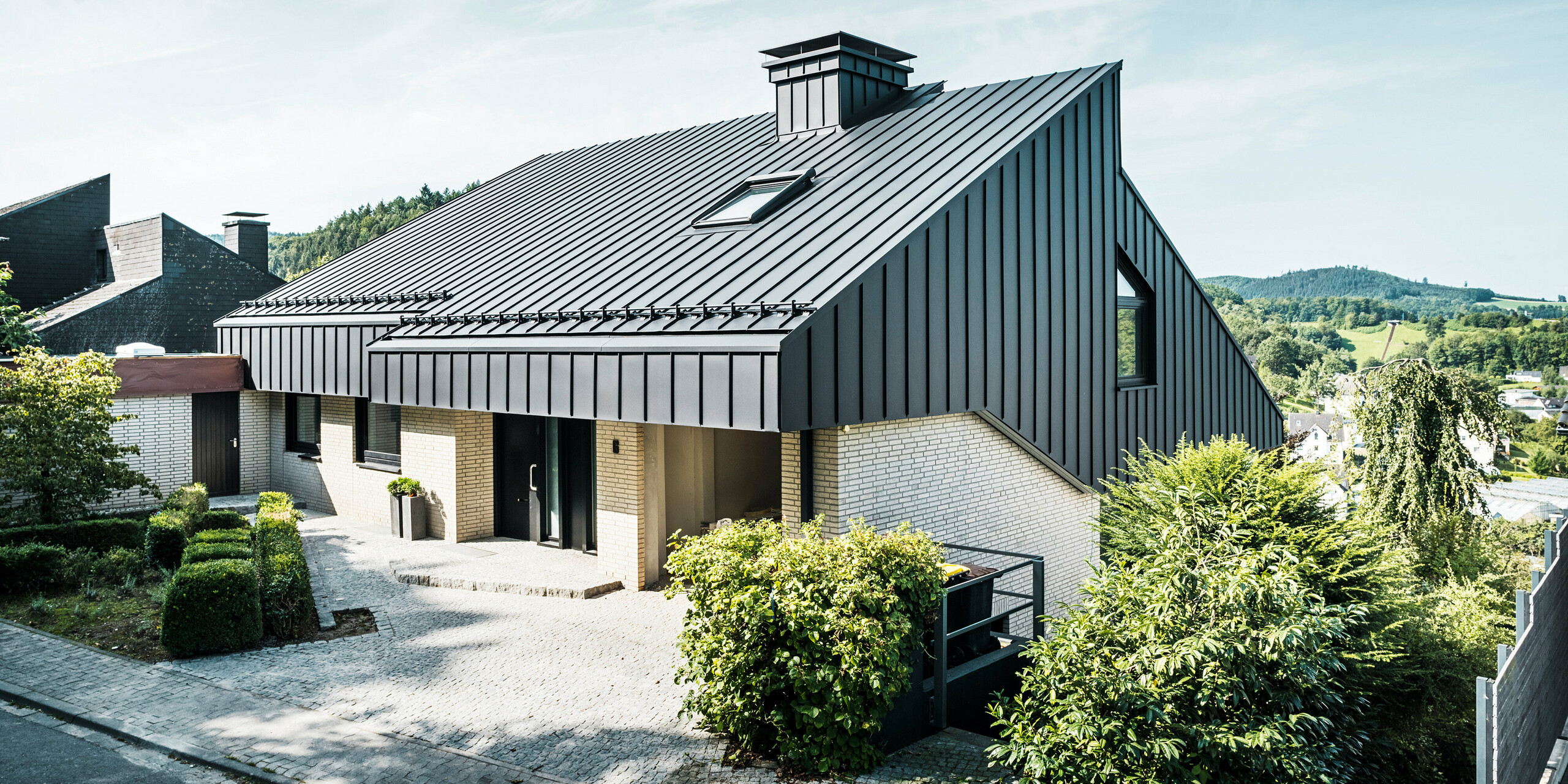 Moderne Architektur eines Einfamilienhauses in Meschede, Deutschland, mit einem eleganten, versetzten Pultdach aus PREFALZ P.10 schwarz. Die Aluminiumfassade harmoniert mit den klaren Linien des Gebäudes, während die Umgebung mit grünen Hecken und Pflanzen das Gesamtbild abrundet. 