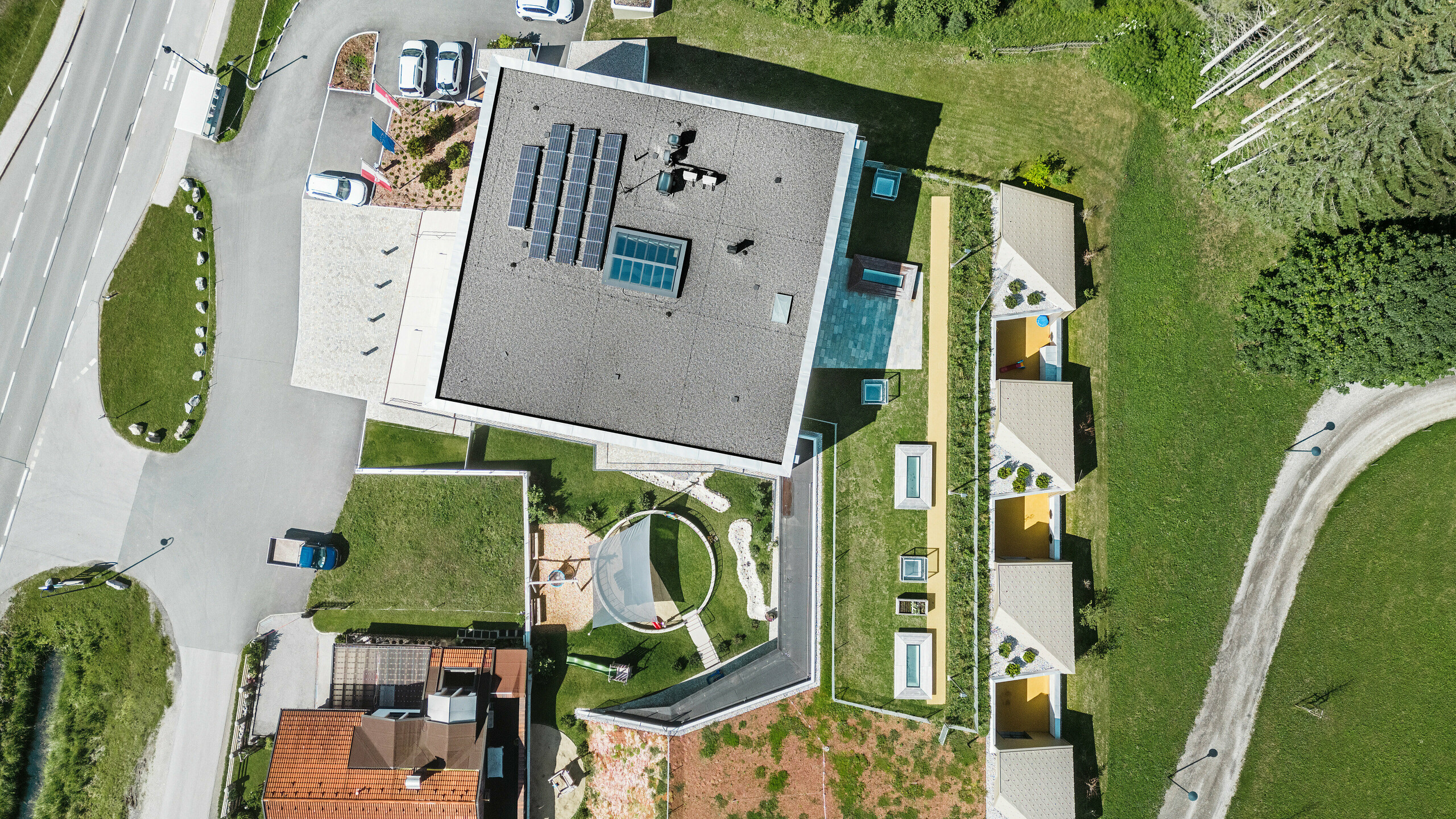 Pohľad z vtáčej perspektívy na vzdelávacie centrum Gerlos so súborom moderných a tradičných budov. Hlavná budova sa prezentuje sivou šikmou strechou vybavenou solárnymi panelmi a je obklopená ďalšími budovami so žltými fasádami a zelenými strechami. V pozadí je vidieť malebnú zalesnenú alpskú krajinu, zatiaľ čo v popredí vidieť zaparkované autá a kľukatú cestu.