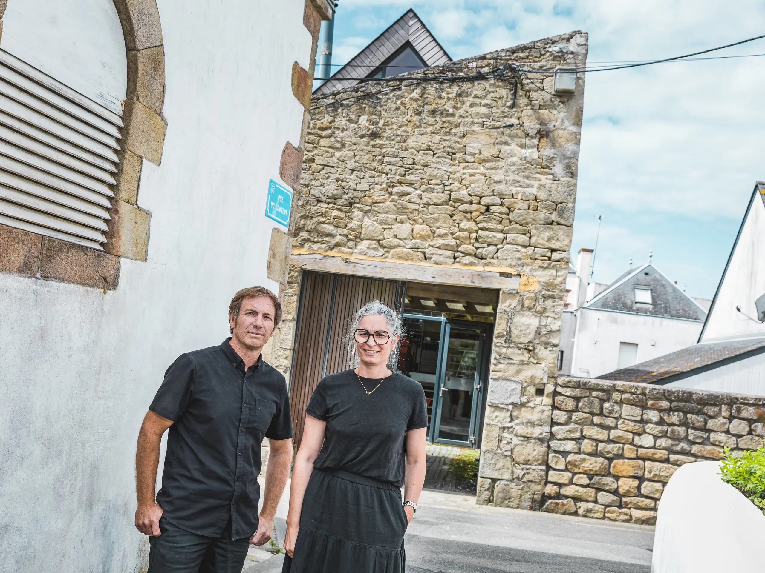 V úzkej bretónskej uličke stoja obaja architekti Anne-Charlotte a Jean-François Riguidelovci pred výraznou fasádou z prírodného kameňa. Obaja sú oblečení celí v čiernom. Pôsobivý kontrast medzi tradičným murivom, bielymi fasádami domov a modernou strešnou architektúrou upriamuje pozornosť na vydarené prepojenie regionálnej stavebnej kultúry so súčasným dizajnom.