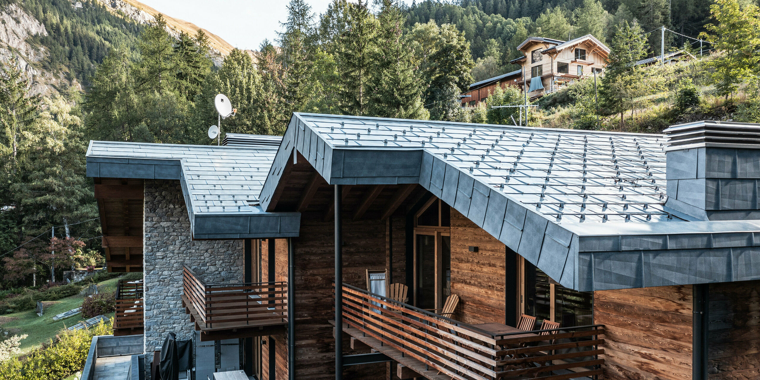 Komplexná vila v Courmayeur s drevenými fasádami a hliníkovými strešnými panelmi PREFA FX.12 vo farbe P.10 kamenná šedá. Architektúra kombinuje moderné a tradičné prvky, ako je murivo z prírodného kameňa a viacposchodovej drevenej konštrukcie, doplnené štýlovými hliníkovými detailmi. Niekoľko balkónov a terás poskytuje dostatok vonkajšieho priestoru doplneného sviežou zeleňou a dramatickou horskou krajinou za nimi.