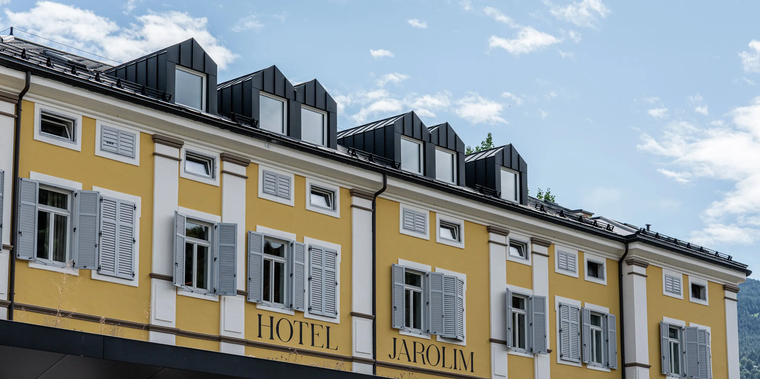 Detailný pohľad na hotel Jarolim v Brixene s modernými strešnými vikiermi, obloženými hliníkom PREFA. Presne vyrobené vikiery a strešné panely R.16 v čiernej farbe P.10 podčiarkujú eleganciu a funkčnosť strechy. Trvanlivá a odolná strecha PREFA sa bezproblémovo začleňuje do historickej architektúry a spája tradíciu s inováciou. Perfektná voľba pre trvácne, estetické a kvalitné strešné riešenia.