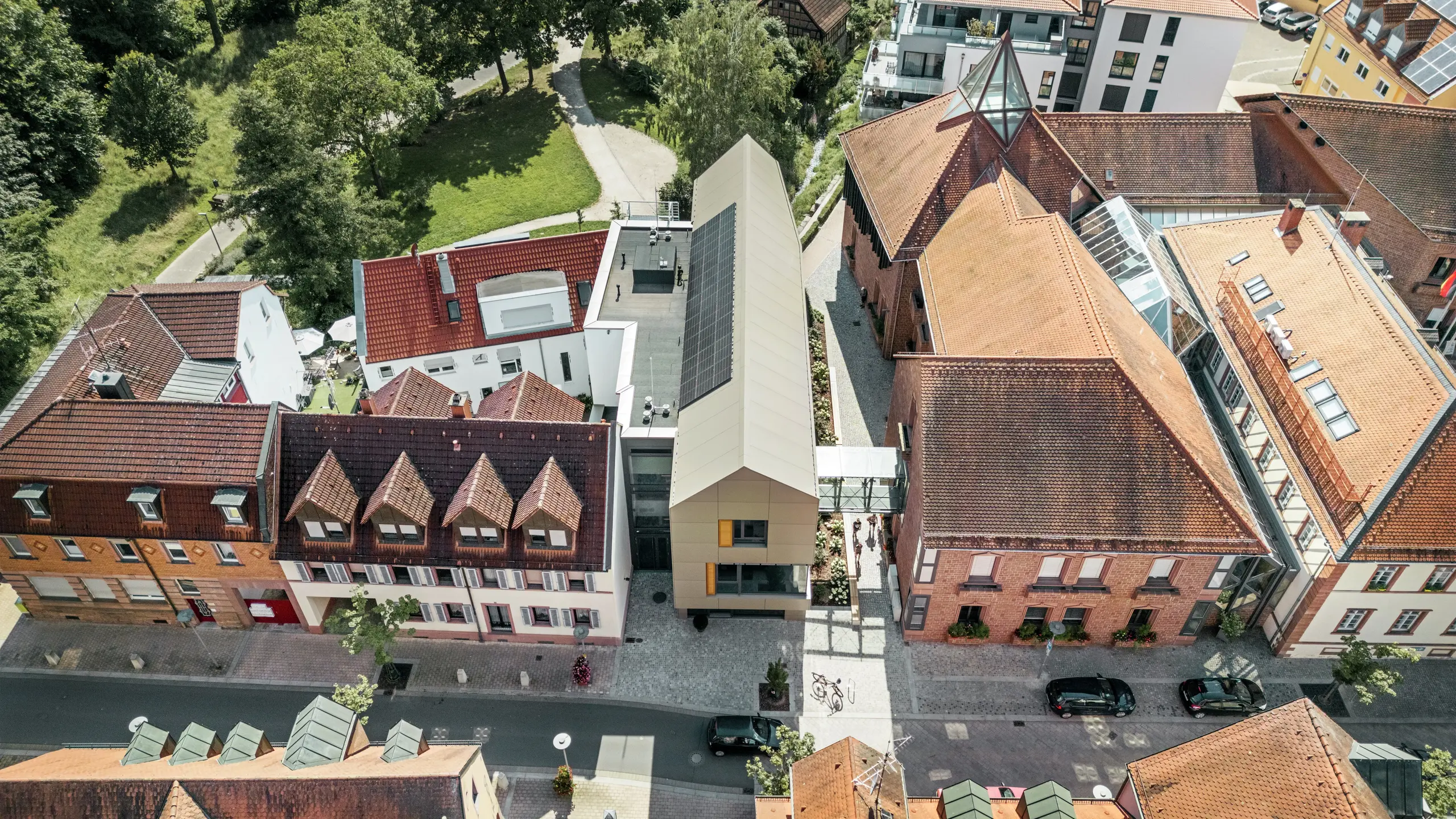 Radnica v nemeckom Alzenau sa nachádza v centre historického mestského prostredia, obklopená tradičnými budovami s červenými škridlovými strechami. V kontraste s nimi stojí moderná radnica, ktorej fasáda je obložená hliníkovými kompozitnými panelmi PREFA v elegantnom bronzovom odtieni. Inovatívna architektúra sa jasne odlišuje od susedných klasických budov, ale napriek tomu sa harmonicky začleňuje do mestského prostredia.