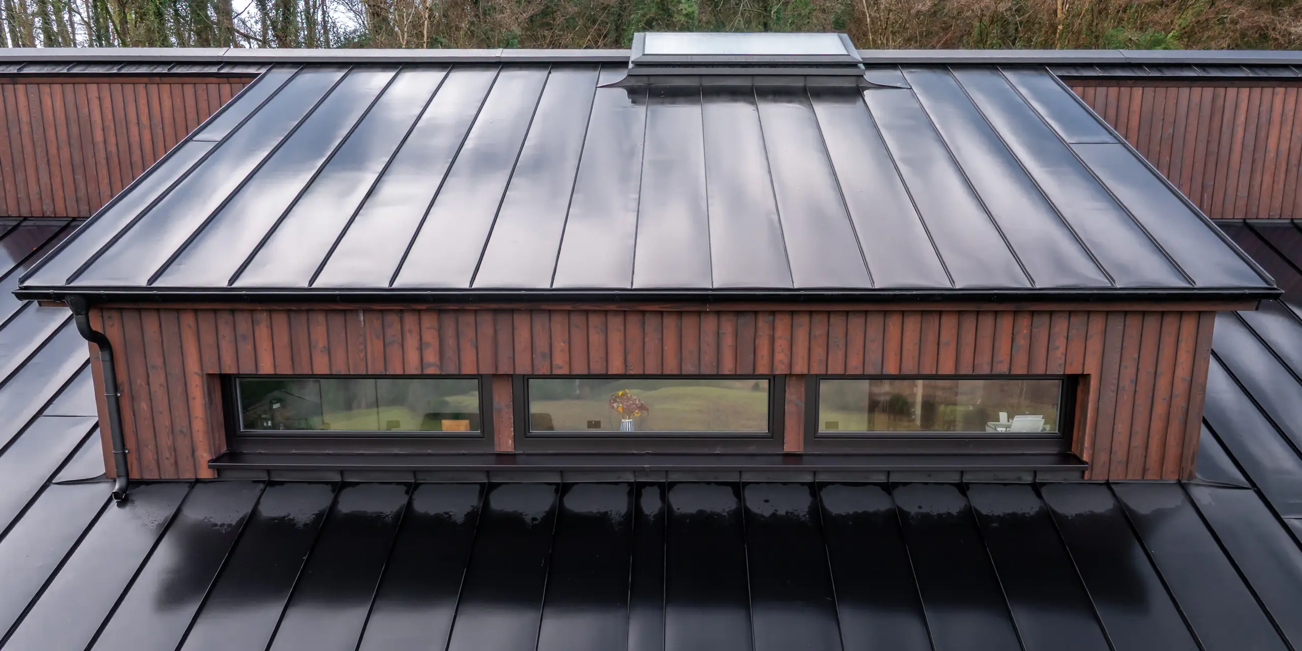 Draufsicht auf die Gaube des House of Holywell in Sligo: Die klar gegliederten Linien des PREFALZ Aluminiumdachs in P.10 Schwarz führen den Blick entlang der präzisen Stehfalze und betonen die reduzierte Formensprache. Die glatte, leicht spiegelnde Oberfläche hebt die hochwertige Verarbeitung hervor und setzt einen spannenden Kontrast zur warmen Holzfassade. Das PREFA Dachsystem überzeugt durch seine Langlebigkeit, Korrosionsbeständigkeit und zuverlässigen Schutz bei wechselhaften Witterungsbedingungen.