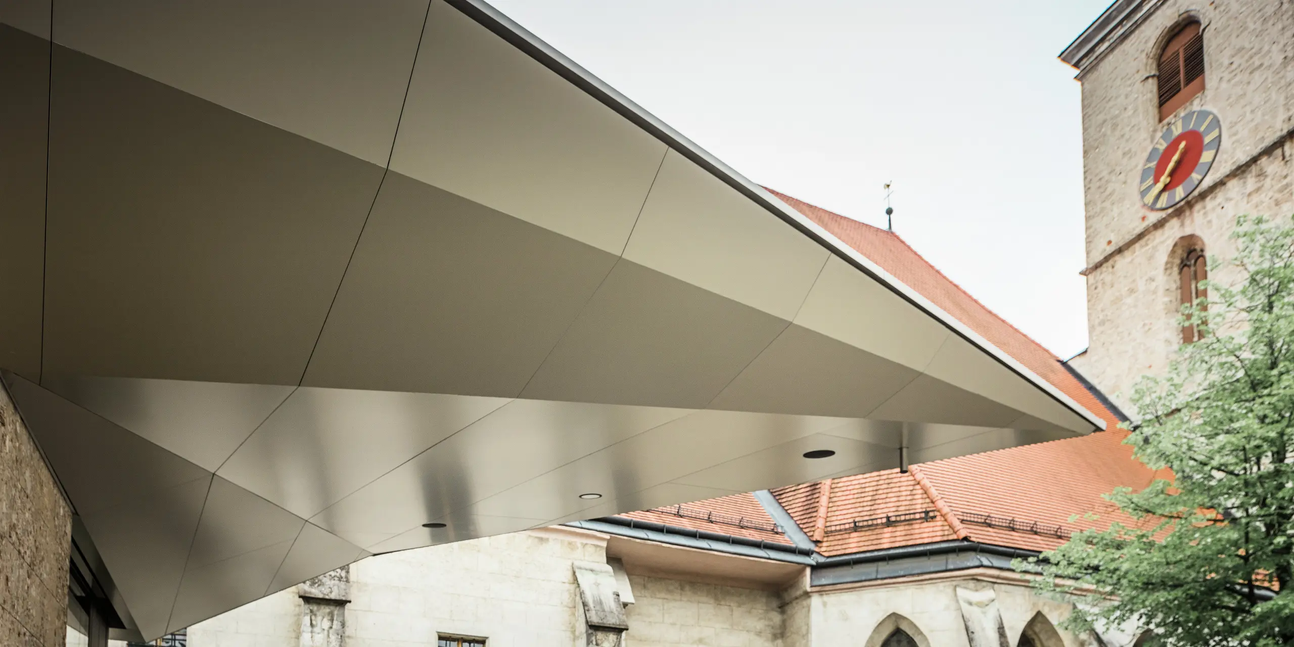 Dynamisch gespannte Dachfläche mit scharfkantiger Linienführung: PREFA Aluminium Verbundplatten ermöglichen diese präzise, fugenarme Gestaltung, die dem Eingangsbereich Leichtigkeit und Modernität verleiht - bei gleichzeitig hoher Witterungsbeständigkeit. Im Hintergrund ist die historische Kirche aus dem 14. Jahrhundert zu erkennen.