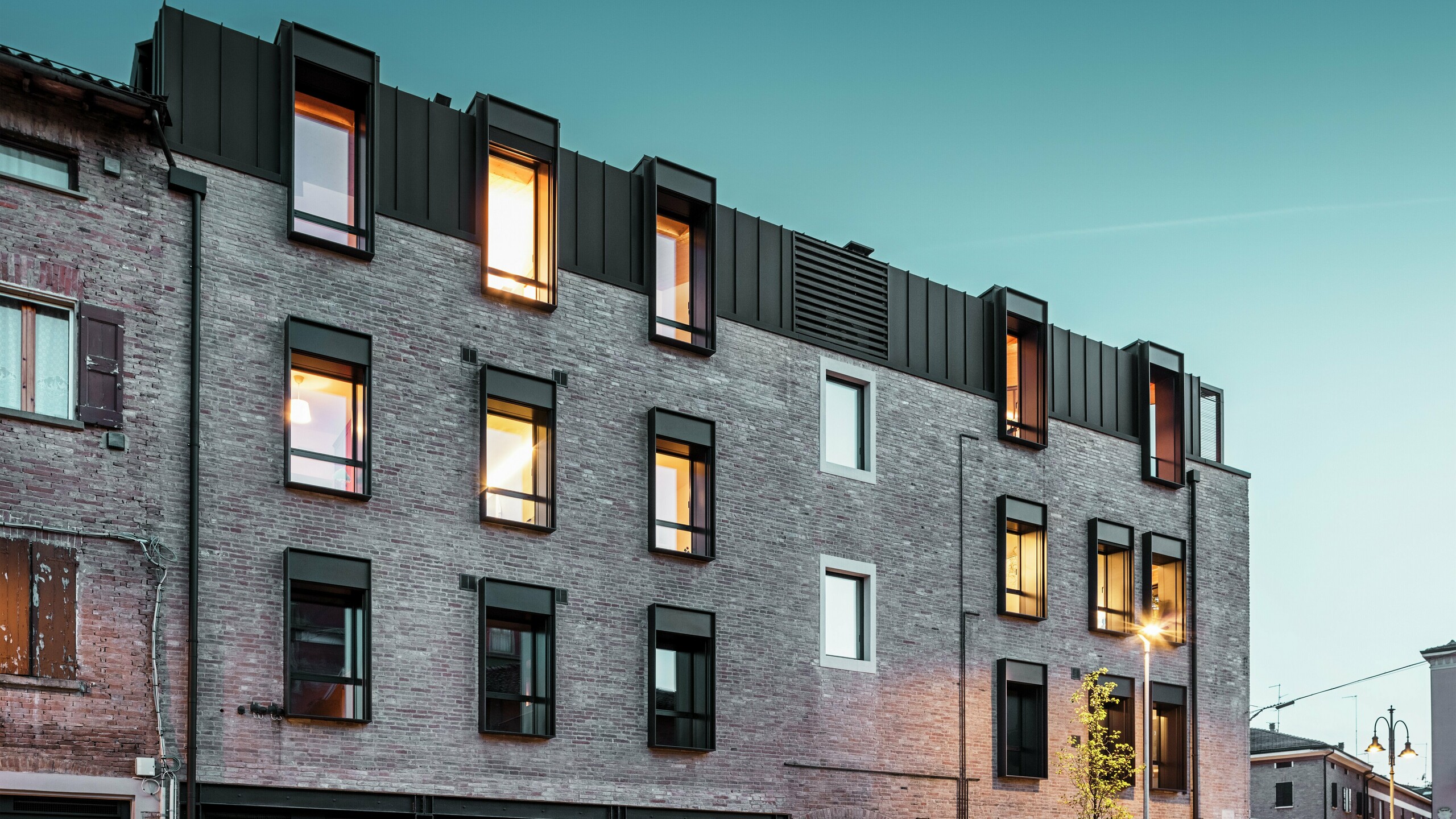 Mestská tehlová budova v Taliansku s modernou hliníkovou strechou PREFALZ v odtieni P.10 orieškovohnedá, osvetlenými oknami a okolitou historickou architektúrou za súmraku.