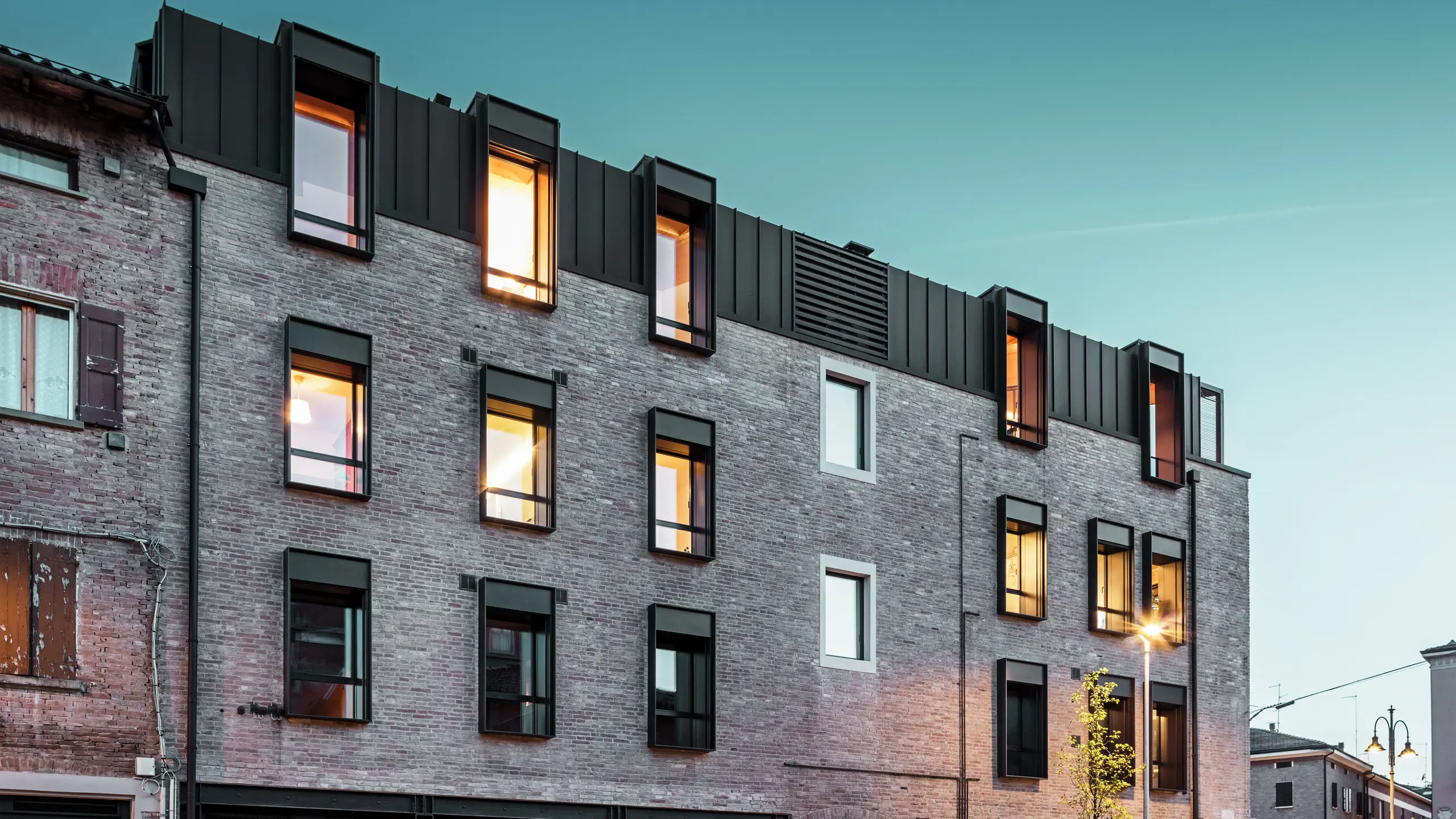 Mestská tehlová budova v Taliansku s modernou hliníkovou strechou PREFALZ v odtieni P.10 orieškovohnedá, osvetlenými oknami a okolitou historickou architektúrou za súmraku.