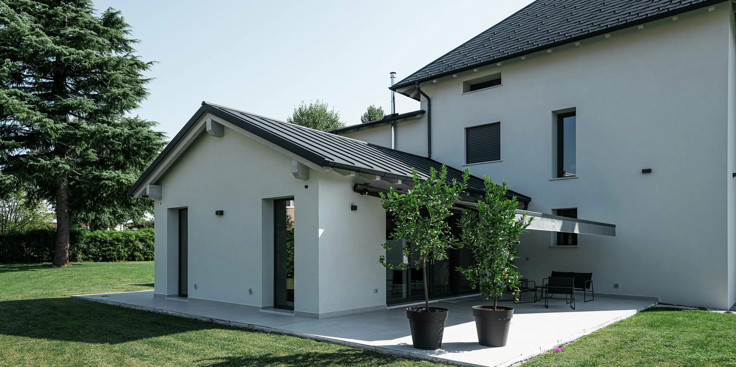 Detailný pohľad na prístavbu moderného rodinného domu v Marano Vicentino v Taliansku s dvojitou stojatou falcovou krytinou PREFA v antracitovej farbe P.10. Elegantná hliníková strecha harmonicky dopĺňa svetlú fasádu a zaisťuje trvácne a odolné riešenie voči poveternostným vplyvom. Priľahlá terasa s ochrannou clonou proti slnku vytvára príjemný vonkajší priestor v záhrade.