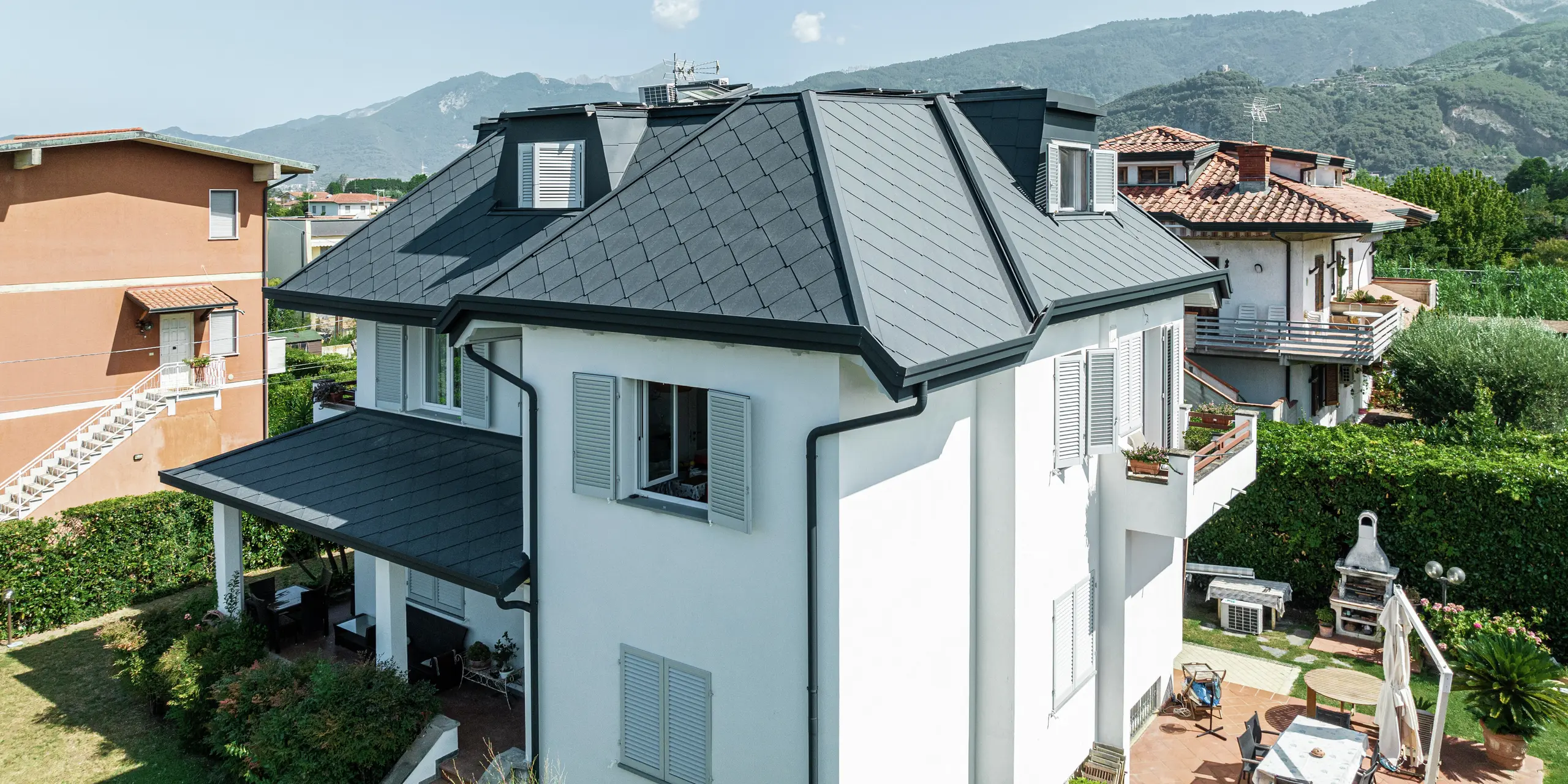 Detailný záber na elegantnú strechu rodinného domu v Taliansku, pokrytú strešnými šablónami PREFA 44 × 44 vo farbe P.10 kamenná šedá. Čisté línie a vysokokvalitná hliníková strešná krytina podčiarkujú moderný dizajn rodinného domu v Montignoso. Vďaka robustným materiálom PREFA je strecha nielen vizuálne príťažlivá, ale aj mimoriadne odolná voči búrkam, korózii a UV žiareniu. Strešný plášť tmavej farby vytvára štýlový kontrast k svetlej fasáde a harmonicky zapadá do okolia