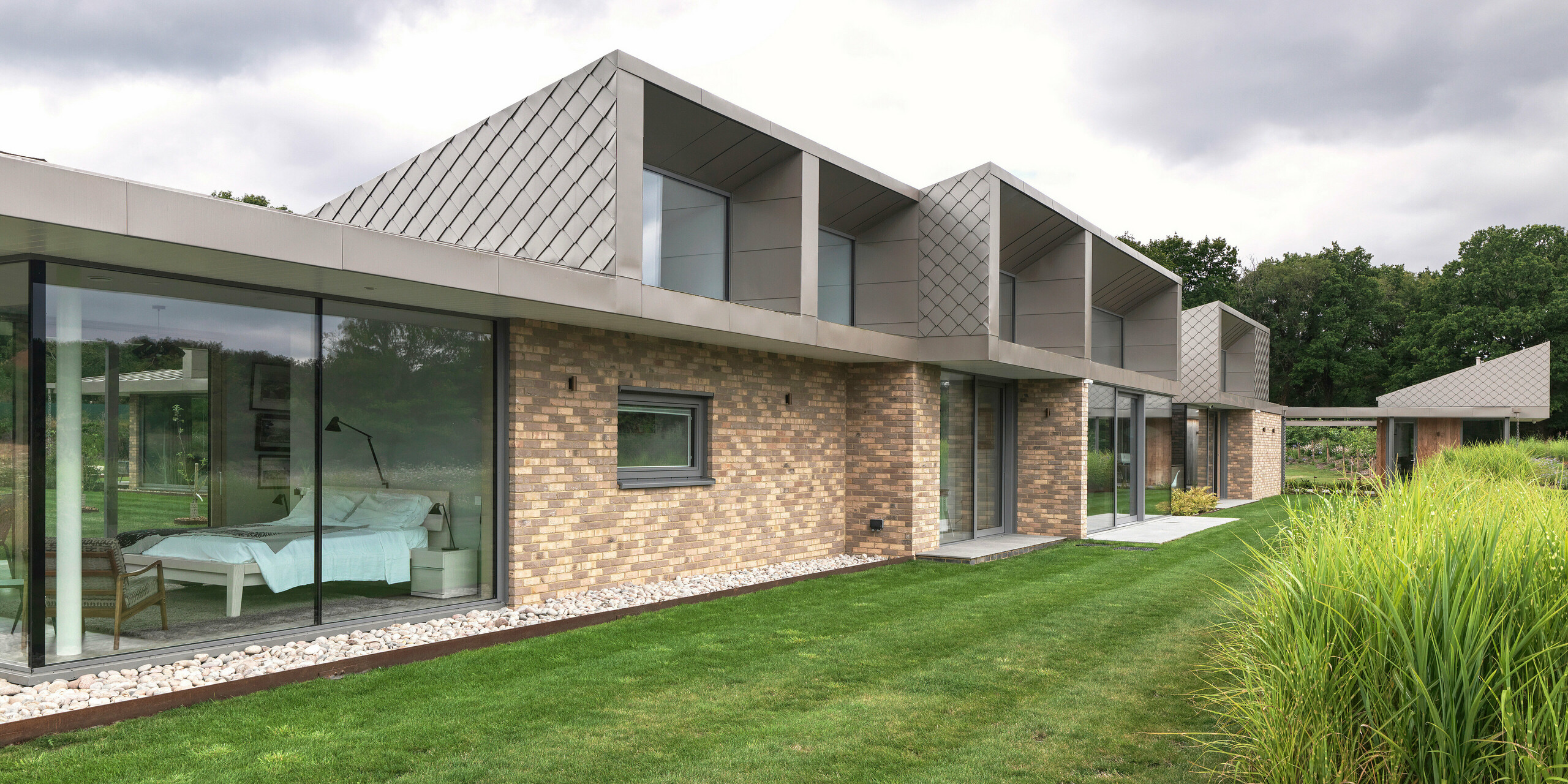 Bočný pohľad na moderný obytný dom v West Sussex s výraznými nadstavbami na poschodí, obloženými PREFA fasádnymi šablónami 20 × 20 v P.10 bronzovej farbe. Ploché sklonené hliníkové strechy PREFALZ tvoria elegantný a odolný proti poveternostným vplyvom povrch. Veľkoplošné zasklenia na prízemí plynule spájajú vnútorný a vonkajší priestor a dodávajú budove kvalitný, nadčasový vzhľad.