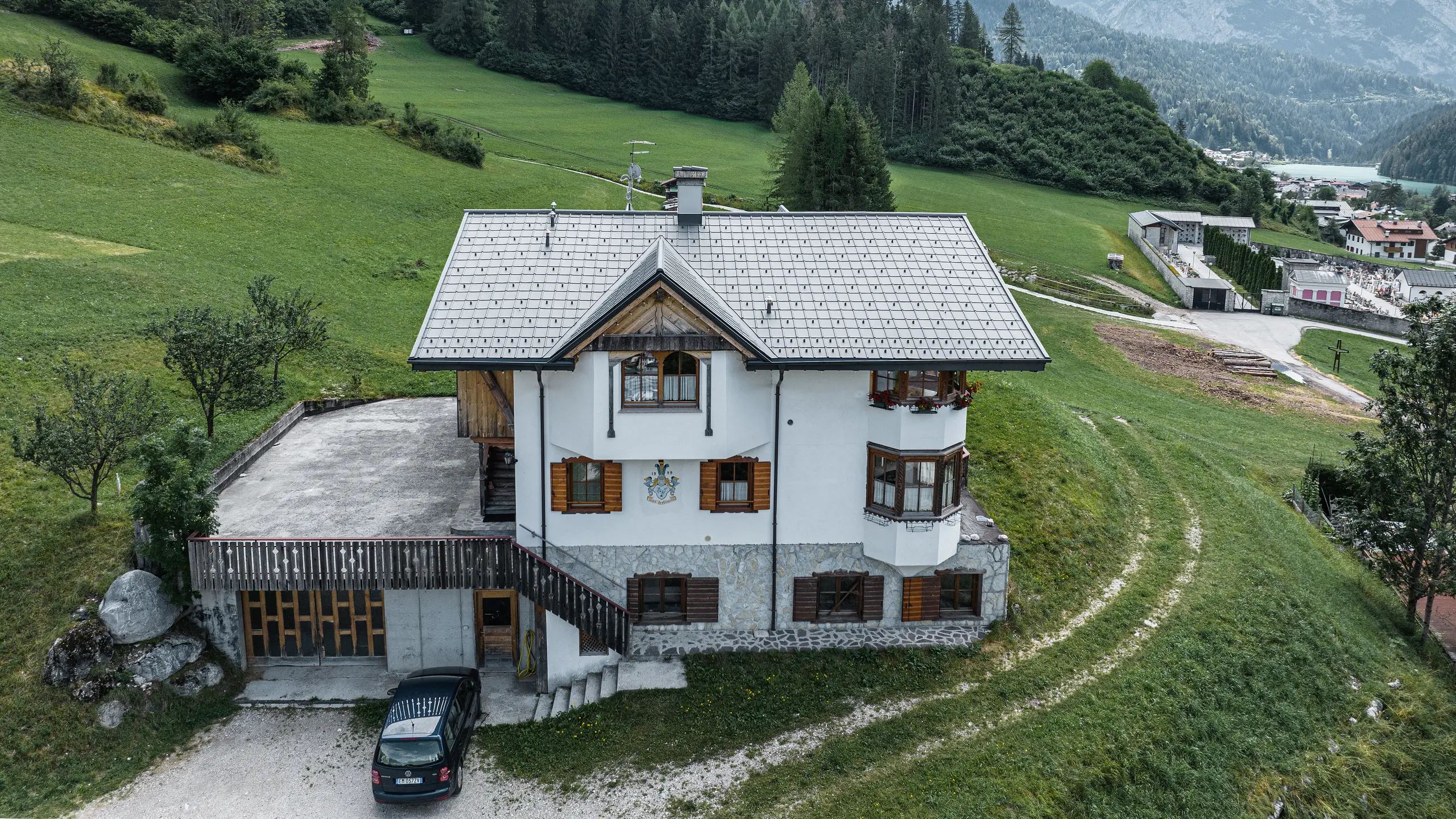 Pohľad z vtáčej perspektívy na vchod do tradičnej horskej chaty v Auronzo di Cadore po rekonštrukcii strechy, na ktorej sú škridly PREFA vo farbe P.10 svetlošedá. Budova s charakteristickým alpským šarmom a modernou strechou je v harmónii so sviežou zelenou krajinou a pôsobivou horskou panorámou v pozadí, čo dokazuje úspešné spojenie funkčnosti a regionálnej architektúry.