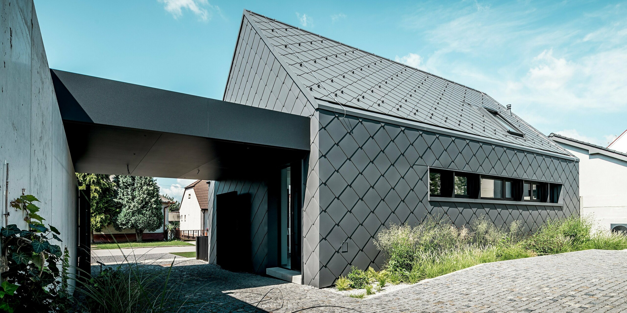 Na obrázku je zachytená moderná architektúra rodinného domu v Trnave, kompletne obložená strešnými a fasádnymi šablónami PREFA 44 × 44 v odtieni P.10 tmavošedá. Súvislá šablónová štruktúra zdôrazňuje čisté geometrické línie budovy a dodáva jej elegantný, monolitický vzhľad. Trvanlivé hliníkové produkty poskytujú dlhodobú ochranu pred poveternostnými vplyvmi a zaisťujú kvalitný plášť budovy s nízkymi nárokmi na údržbu. Svetlom preplnená predná časť a okolité zeleň vytvárajú prirodzený kontrast k výraznej fasáde PREFA.