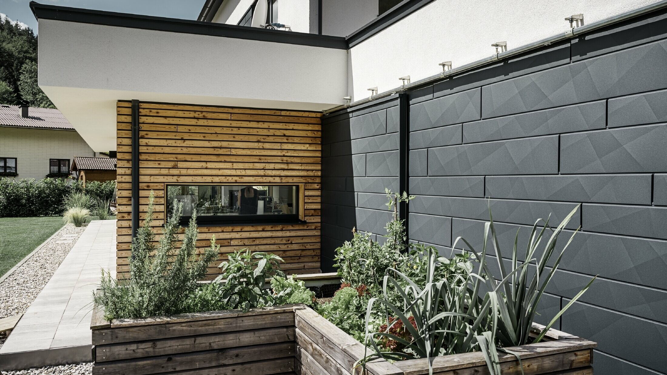 Tento obrázok ukazuje detailný záber rodinného domu so zameraním na fasádny obklad s PREFA Siding.X vo farbe P.10 Antracit. Tmavé fasádne panely s výrazným zvrásneným vzhľadom poskytujú príťažlivý vizuálny kontrast k živým, drevom obloženým plochám a okolitým zeleným priestranstvám. Vyvýšené záhony vysadené rôznymi rastlinami a kríkmi sú umiestnené pred hliníkovou fasádou, čo podčiarkuje prirodzený a udržateľný vzhľad domu. Kombinácia moderných materiálov a začlenenie do záhradnej plochy sprostredkúva obraz súčasnej architektúry, ktorá harmonicky zapadá do svojho okolia.