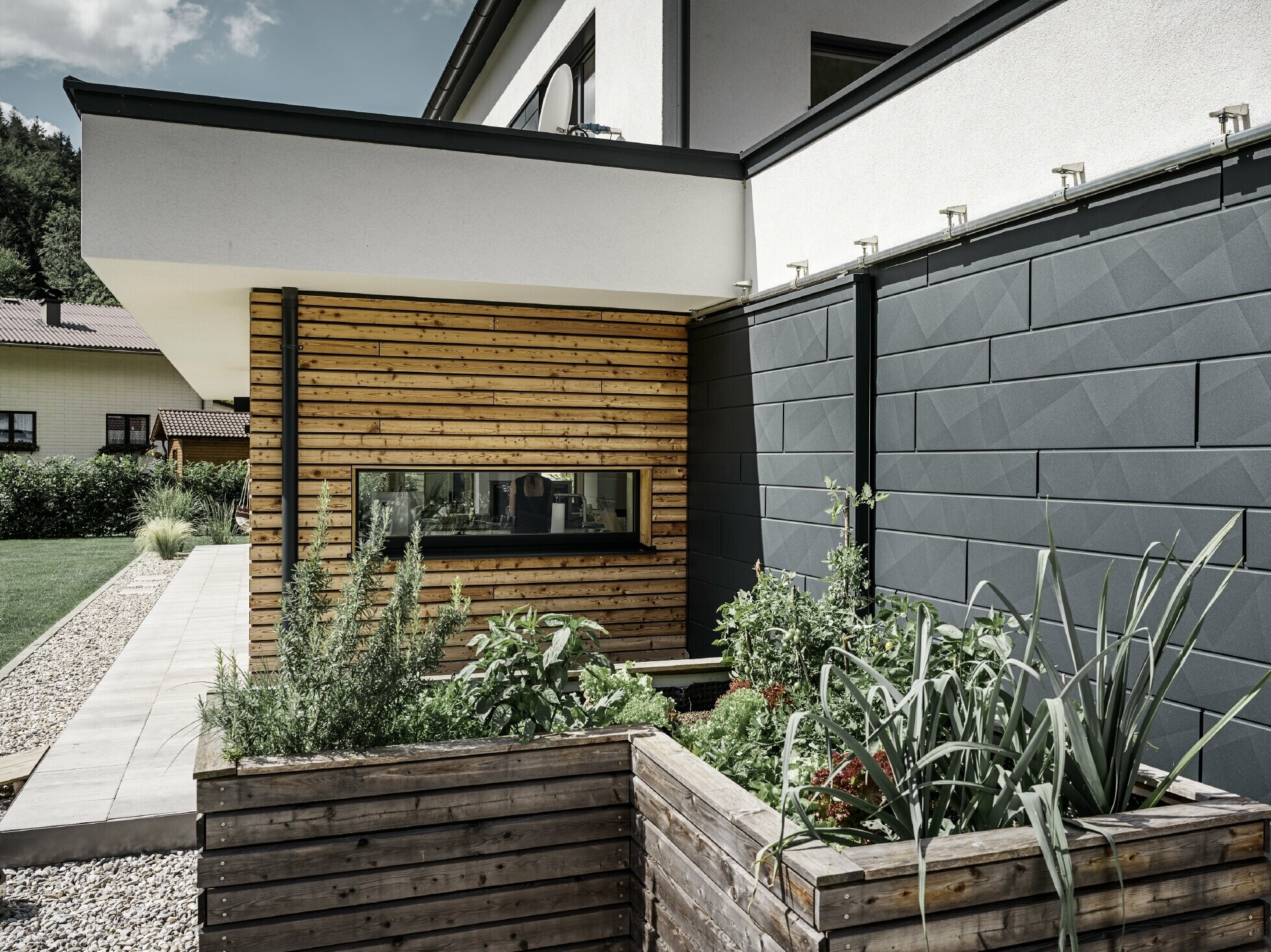 Tento obrázok ukazuje detailný záber rodinného domu so zameraním na fasádny obklad s PREFA Siding.X vo farbe P.10 Antracit. Tmavé fasádne panely s výrazným zvrásneným vzhľadom poskytujú príťažlivý vizuálny kontrast k živým, drevom obloženým plochám a okolitým zeleným priestranstvám. Vyvýšené záhony vysadené rôznymi rastlinami a kríkmi sú umiestnené pred hliníkovou fasádou, čo podčiarkuje prirodzený a udržateľný vzhľad domu. Kombinácia moderných materiálov a začlenenie do záhradnej plochy sprostredkúva obraz súčasnej architektúry, ktorá harmonicky zapadá do svojho okolia.