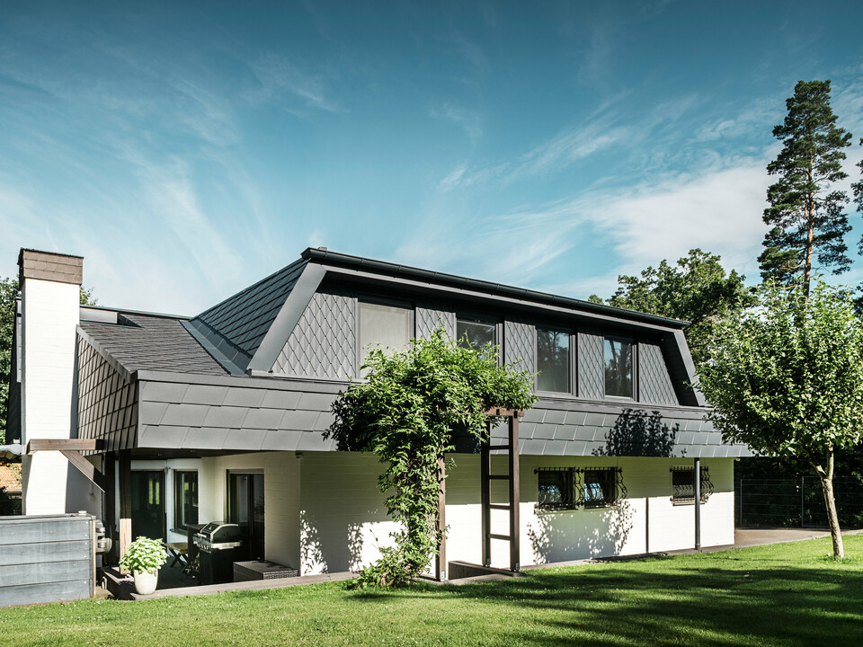 Bočný pohľad na rodinný dom v Mechernich so strešnými šindľami PREFA DS.19 a malými strešnými šablónami v antracitovej farbe P.10. Trapézové vikiere sú opláštené strešnými šablónami z robustného plechu, zatiaľ čo strecha je pokrytá šindľom. Modernú architektúru odolnú voči poveternostným vplyvom dopĺňa zelená pergola a stromy v záhradnej časti.