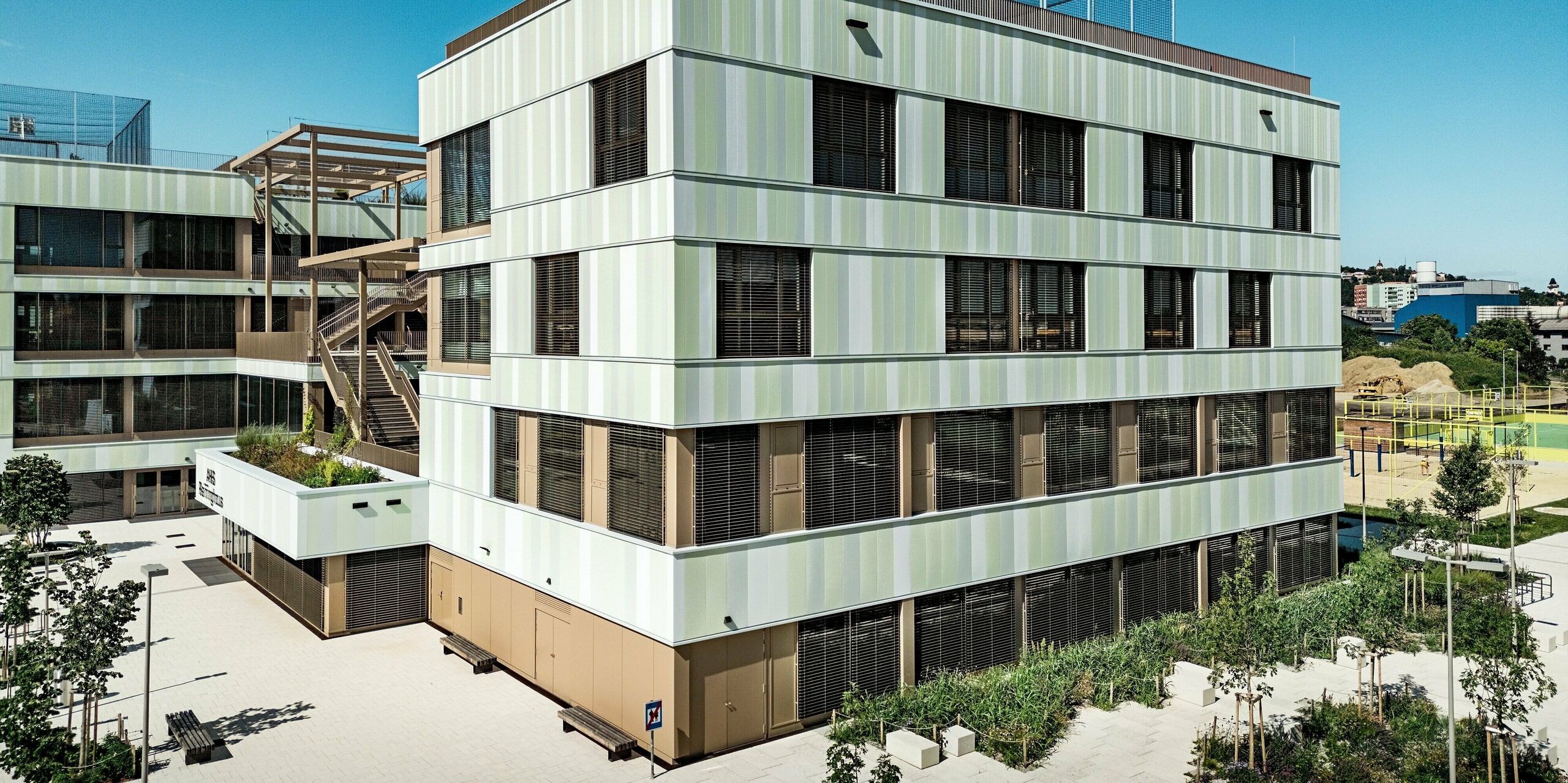 Das Bild zeigt die beeindruckende Fassade der AHS Reininghaus in Graz. Sie sticht durch ihre modernen PREFA Sidings in Sonderfarben hervor. Die vertikal verlaufenden Aluminiumprofile sorgen für eine lebendige Struktur. Besonders auffällig sind die bräunlichen Fensterrahmen und Jalousien, die sowohl funktionale als auch ästhetische Akzente setzen. Die PREFA Sidings machen die Fassade besonders widerstandsfähig und pflegeleicht. Sie schützen das Gebäude vor Witterungseinflüssen und werten es gleichzeitig optisch auf. Der angrenzende Außenbereich mit grünen Pflanzen und Sitzbänken ergänzt das Gesamtbild und sorgt für eine ansprechende Atmosphäre.