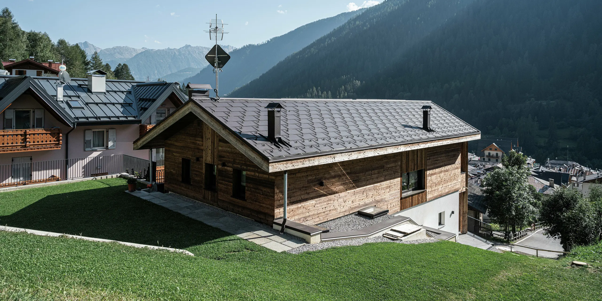 Zadná časť modernej horskej chaty v Cogolo v talianskom údolí Val di Sol s odolnými strešnými panelmi PREFA R.16 v hnedej farbe P.10. Robustná hliníková strecha dokonale zapadá do alpskej krajiny a spoľahlivo chráni budovu pred snehom, vetrom a poveternostnými vplyvmi. Jasné línie a maximálna nosnosť robia z tejto strechy ideálne riešenie pre vysokohorské oblasti.