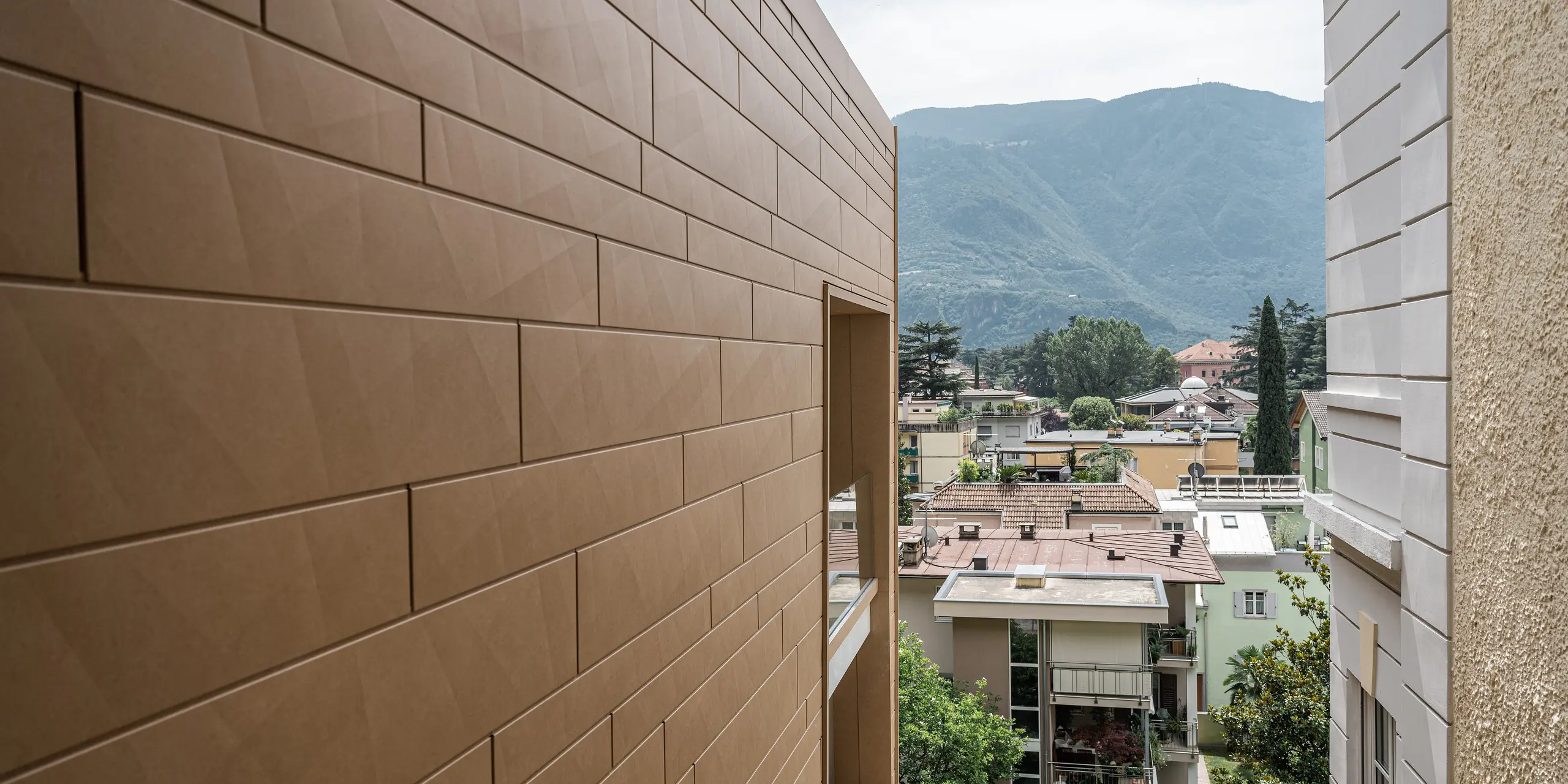 Detailný záber na modernú fasádu PREFA Siding.X v pieskovo hnedej farbe P.10 na vile Laura v Bolzane, Taliansko. Harmonicky spojené hliníkové prvky ponúkajú odolné a estetické riešenie, ktoré elegantne zapadá do malebnej horskej scenérie Južného Tirolska. Fasáda odráža inovácie a kvalitu, ktorými sú hliníkové výrobky PREFA preslávené. Okrem toho hliníkový plášť budovy poskytuje teplý farebný akcent v mestskej krajine.
