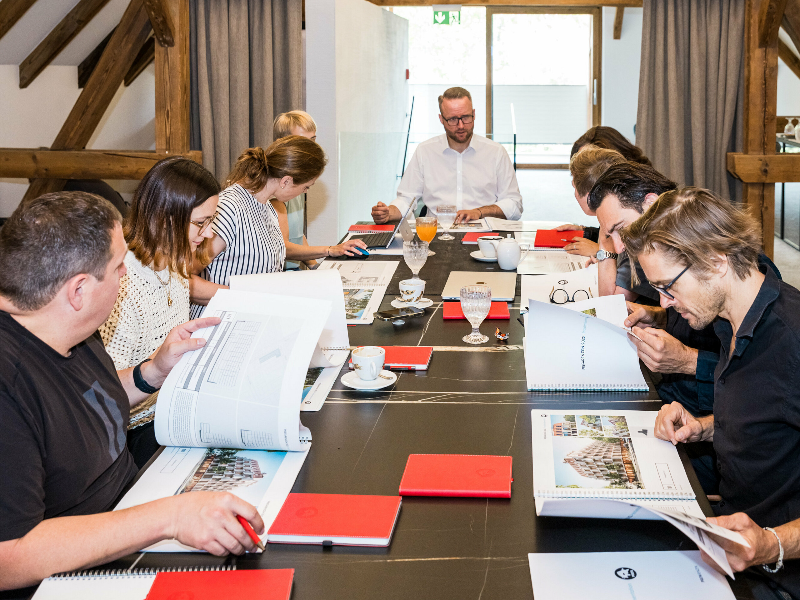 Dialóg expertov PREFARENZEN 2025: Desať architektov a expertov PREFA sedí za konferenčným stolom v sídle vinohradníka v regióne Seewinkel v Burgenlande. Na stole sú vyložené dokumenty a architektonické plány, zatiaľ čo účastníci intenzívne diskutujú a vyberajú dvanásť najzaujímavejších architektonických projektov pre architektonickú platformu PREFA 2025. Diskusia sa zamerala na ekologické a sociálne výzvy, funkčnosť, jednoduchosť a pridanú hodnotu architektúry. Na stole sú rozložené aj šálky na kávu, poháre a červené zápisníky, ktoré podčiarkujú sústredenú pracovnú atmosféru.