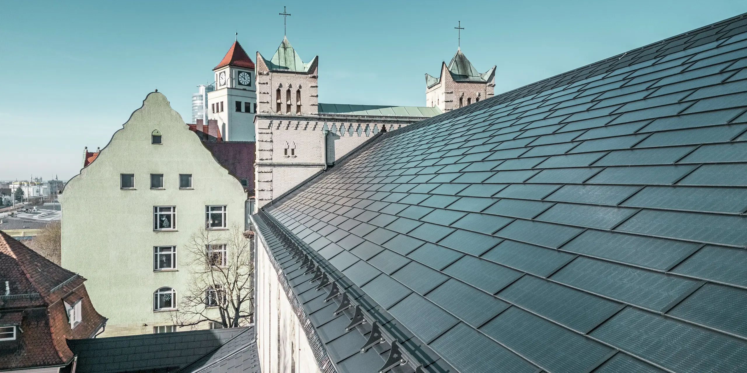 Na detailnom zábere strechy kostola svätého Karola Boromejského v Norimbergu sú vidieť precízne položené integrované solárne panely PREFA v elegantnej čiernej farbe. Dobre integrované solárne moduly harmonicky zapadajú do historickej architektúry a poskytujú udržateľný zdroj energie. V pozadí je vidieť nápadné kostolné veže s klasickými špicatými strechami. Strešné riešenie PREFA z robustného hliníka dokonale spája ochranu pamiatky s obnoviteľnou energiou.