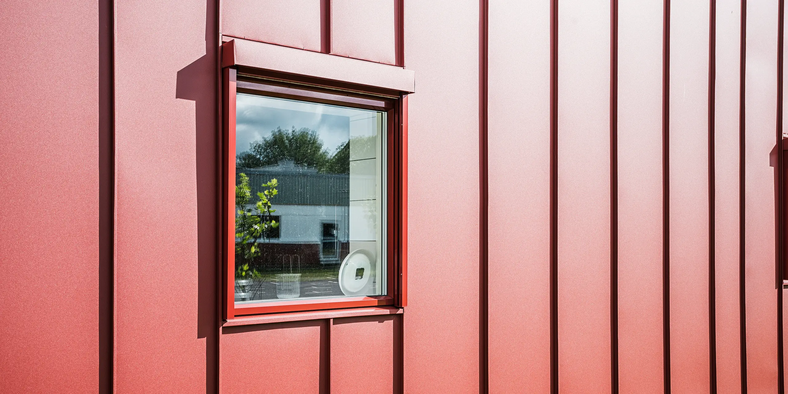 Detailný záber na okno školskej jedálne Gröna Hjärtat v meste Broby vo Švédsku ukazuje detailnú fasádu PREFALZ v tmavočervenej farbe. Precízne spracovanie hliníkového obkladu so stojatou drážkou podčiarkuje vysokú kvalitu a odolnosť obvodového plášťa budovy. Okno je harmonicky začlenené do fasády a odráža okolie, čím zdôrazňuje modernú a jasnú architektúru budovy. Vďaka príťažlivému dizajnu je budova príkladom modernej a udržateľnej výstavby.