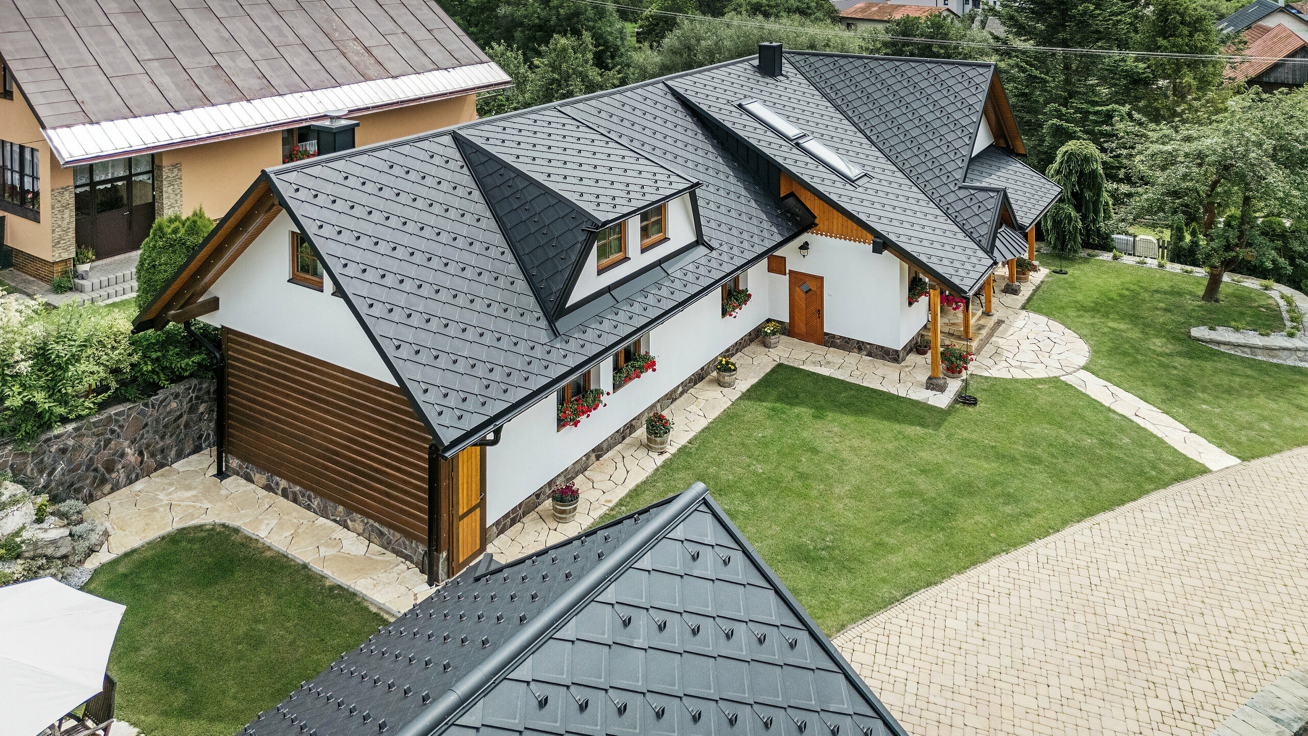 Letecký pohľad na tradičný rodinný dom v Starej Bystrici na Slovensku, pokrytý strešnými šablónami PREFA 29 × 29 vo farbe P.10 čierna. Strecha zaujme elegantným pultovým vzhľadom a harmonickým začlenením vikierov a strešných okien. Okolitá zeleň, udržiavaná záhrada a vydláždená príjazdová cesta podčiarkujú príťažlivý celkový vzhľad budovy.