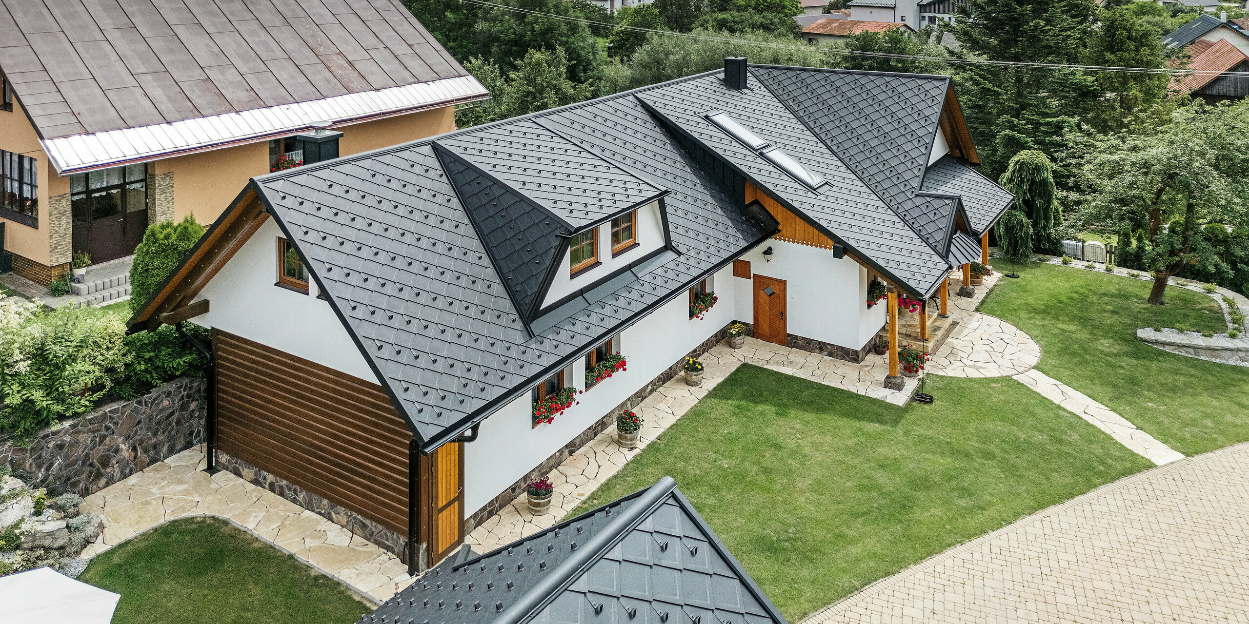 Letecký pohľad na tradičný rodinný dom v Starej Bystrici na Slovensku, pokrytý strešnými šablónami PREFA 29 × 29 vo farbe P.10 čierna. Strecha zaujme elegantným pultovým vzhľadom a harmonickým začlenením vikierov a strešných okien. Okolitá zeleň, udržiavaná záhrada a vydláždená príjazdová cesta podčiarkujú príťažlivý celkový vzhľad budovy.