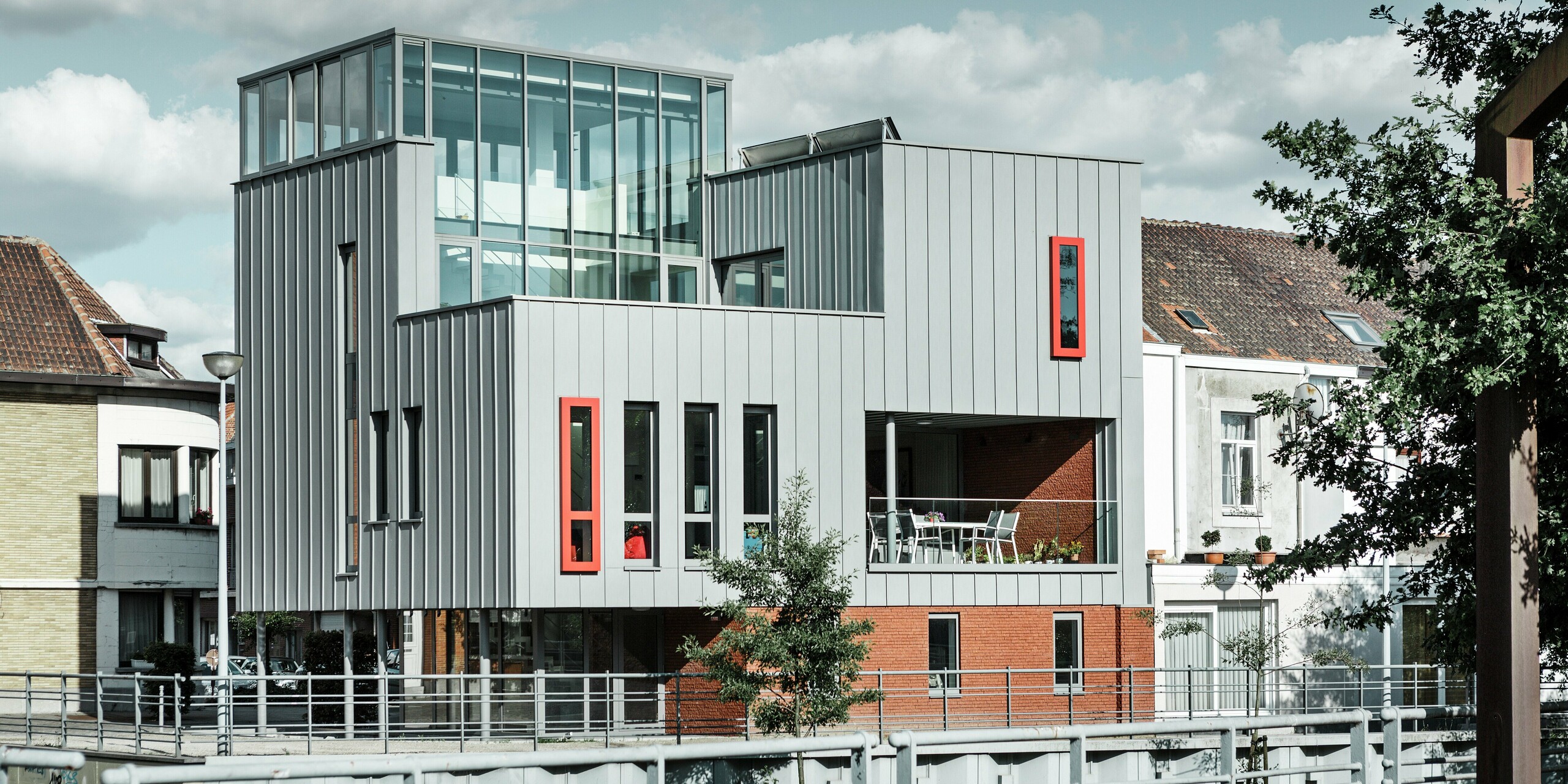 Moderná architektúra na brehu rieky: Obytná a administratívna budova kombinuje elegantnú fasádu PREFALZ v patinovo šedej farbe s nápadnými červenými okennými rámami, ktoré priťahujú pozornosť. Jasne štruktúrovaný vzhľad stojatých falcov sa snúbi s veľkorysými sklenenými plochami a krytou terasou. Hliník ako stavebný materiál tu presviedča svojou odolnosťou voči poveternostným vplyvom, tvarovou stabilitou a nadčasovým dizajnom.  Translated with DeepL.com (free version)