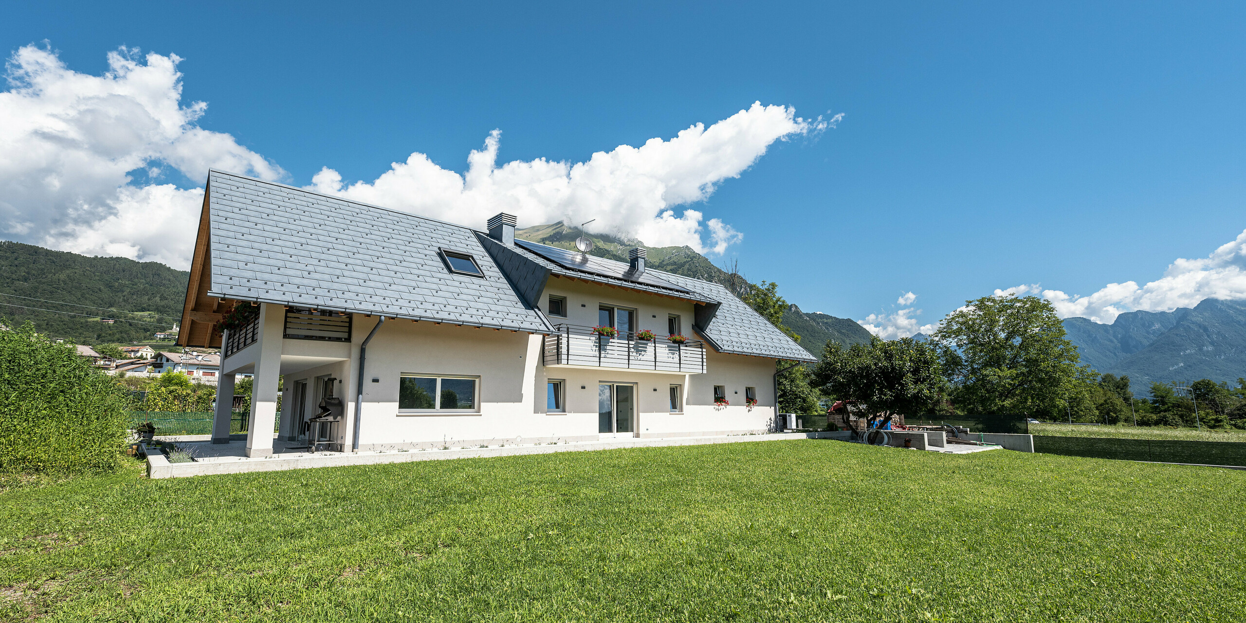  Idylicky umiestnený rodinný dom v talianskom Bellune s robustnou hliníkovou strechou zo strešných panelov PREFA R.16 vo farbe P.10 antracit. S tradičnou bielou omietkou a modernou hliníkovou strechou nehnuteľnosť harmonicky zapadá do malebnej krajiny s úchvatným výhľadom na okolité hory.