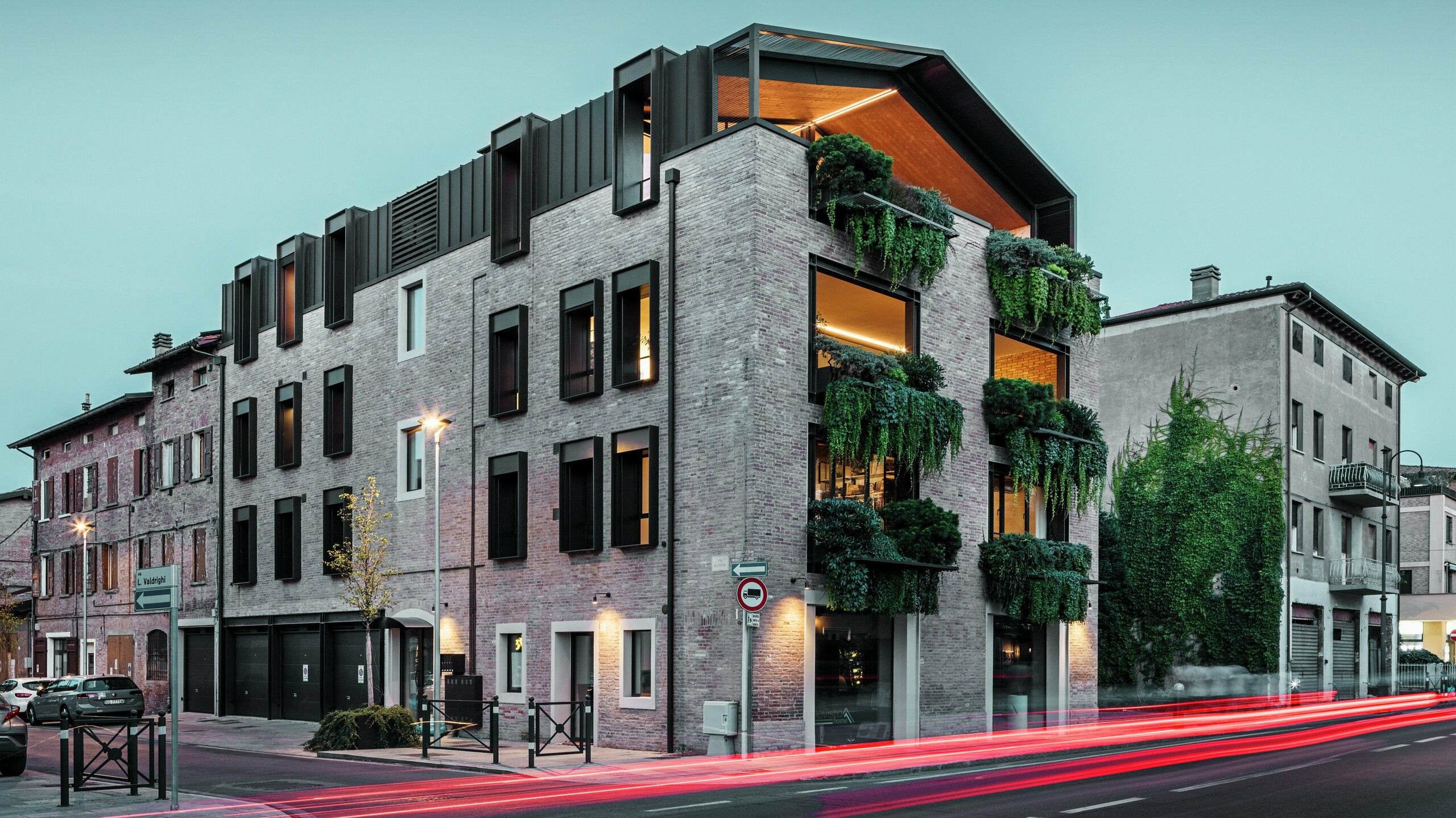 Udržateľná obytná a komerčná budova v Taliansku s hliníkovou strechou PREFALZ v odtieni P.10 orieškovonedá, zelenou tehlovou fasádou a modernou architektúrou v podvečernom prostredí s osvetlenými pouličnými lampami.