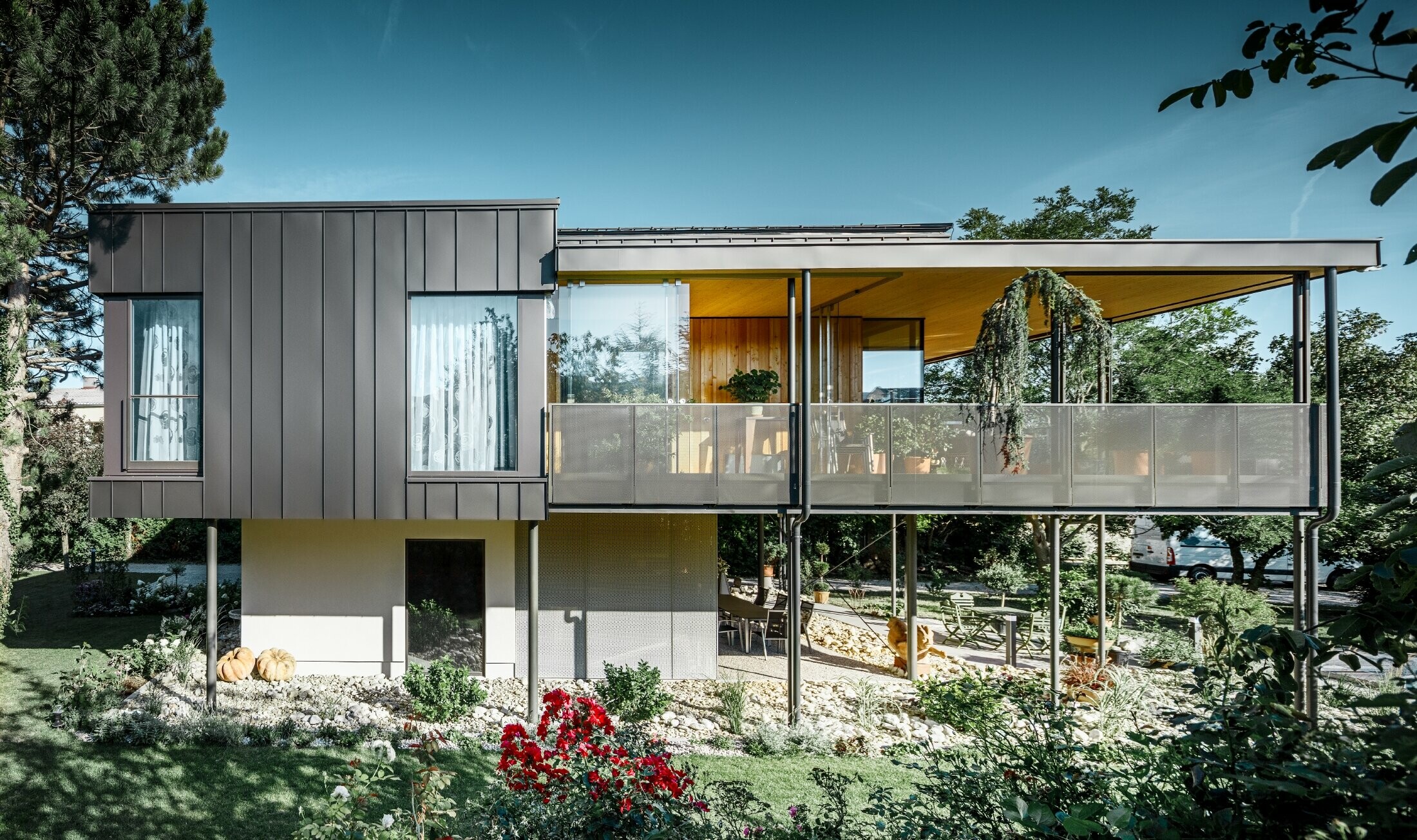 Moderný rodinný dom, podobný drevenému domu obklopenému veľkou záhradou s množstvom stromov, s decentnou uhlovou stojatou drážkou v čiernošedej farbe od fy PREFA