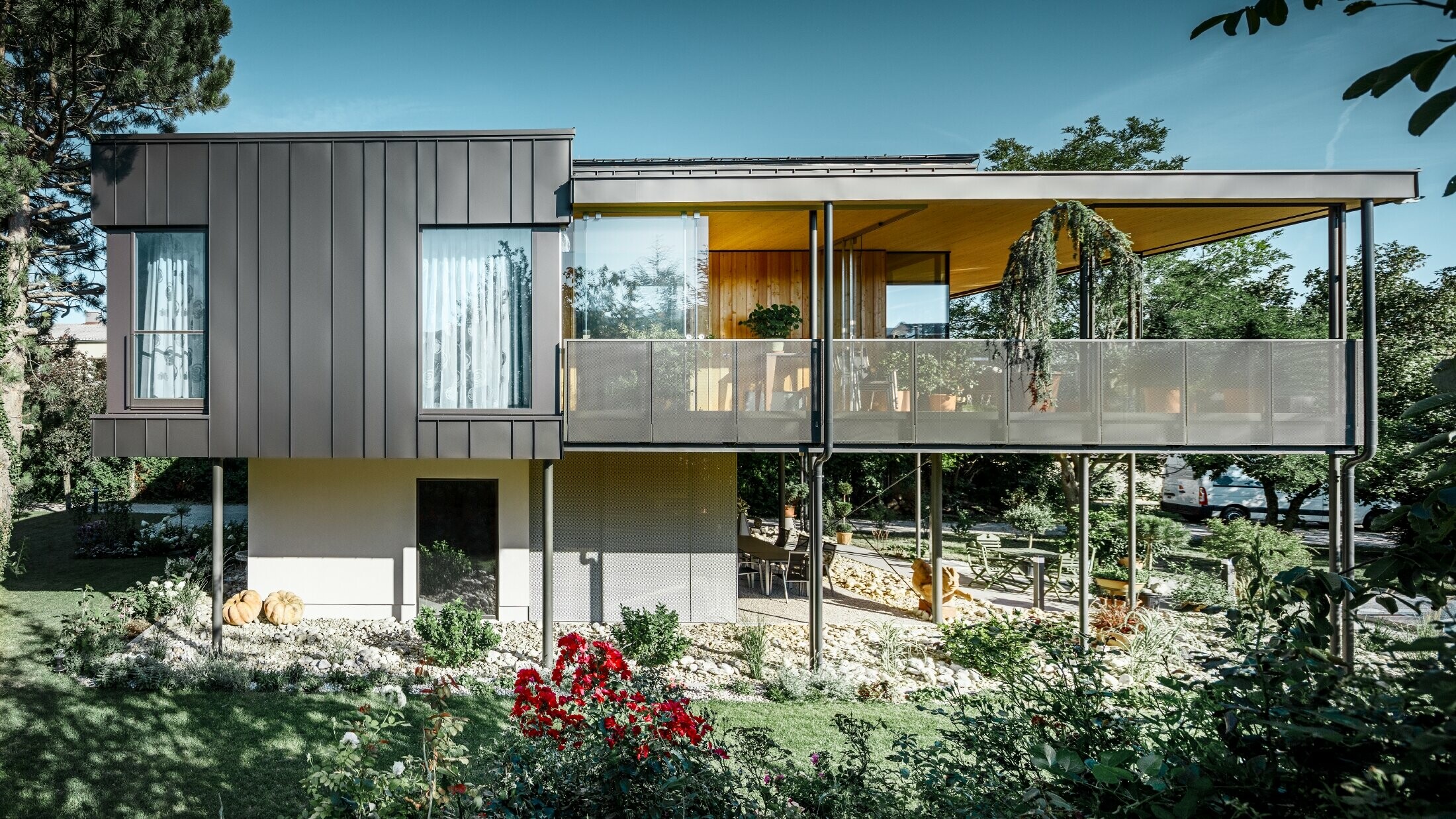 Moderný rodinný dom, podobný drevenému domu obklopenému veľkou záhradou s množstvom stromov, s decentnou uhlovou stojatou drážkou v čiernošedej farbe od fy PREFA