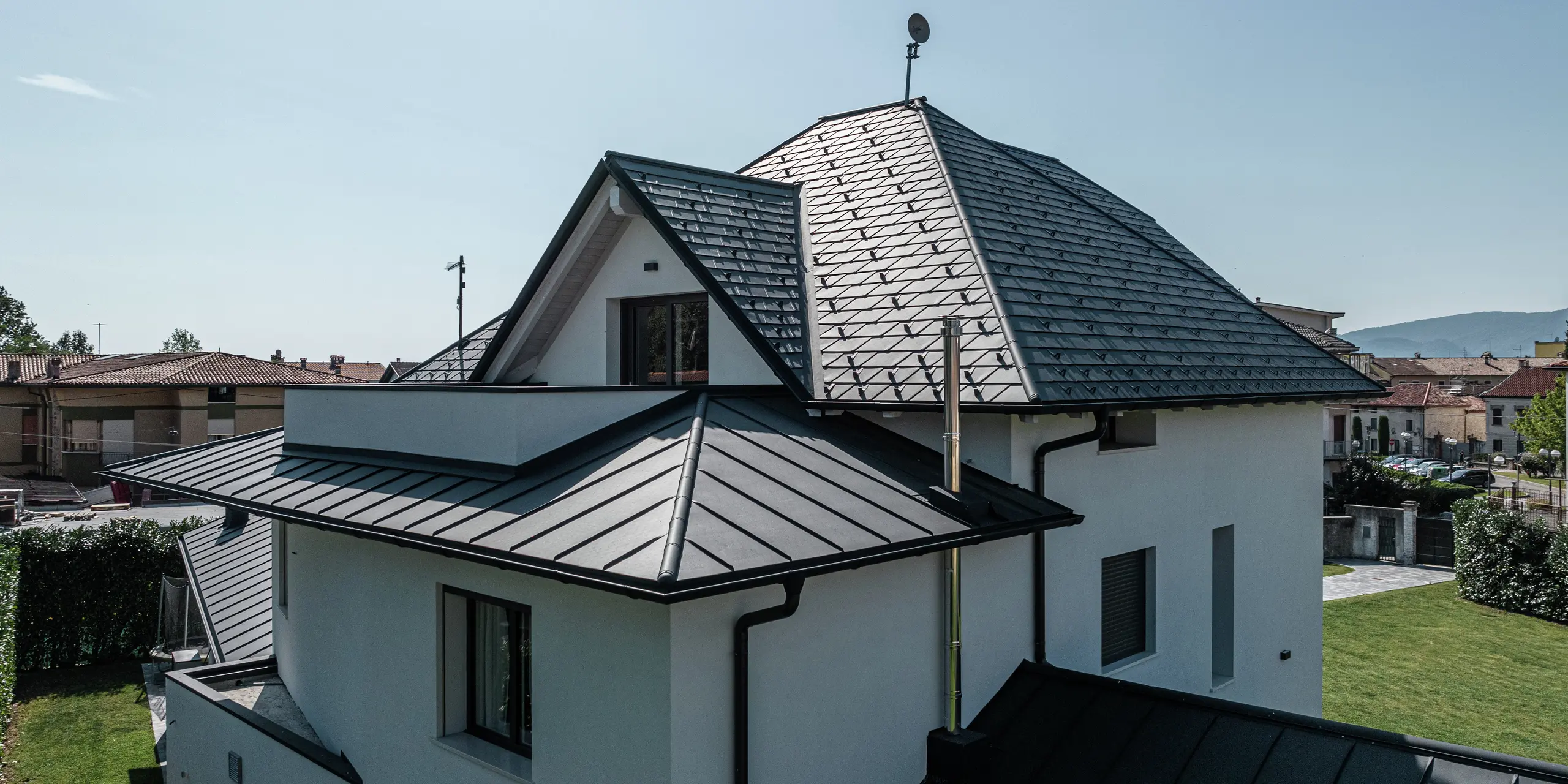 Detailný pohľad na zadnú stranu strechy moderného rodinného domu v Marano Vicentino v Taliansku. Hlavná strecha je pokrytá strešnými šindľami PREFA v antracitovej farbe P.10, zatiaľ čo obe prístavby sú pokryté elegantnou dvojitou stojatou falcovou krytinou z hliníka. Kombinácia vysoko kvalitných hliníkových produktov zaručuje trvácne, odolné voči poveternostným vplyvom a estetické riešenie strechy.