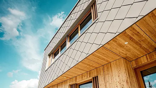 Detailný záber detského zariadenia v Alzenau ukazuje precízne vyhotovenie a vysokokvalitnú kombináciu materiálov z bronzovo sfarbených PREFA fasádnych šablón 44 × 44 a prírodného, neupravovaného dreva. Diagonálna štruktúra hliníkovej fasády vytvára výrazný šupinový vzor, ktorý sa harmonicky prepája s vertikálnymi drevenými lamelami pri oknách a jemne spracovaným dreveným podhľadom. Záber z nadhľadu od zeme (tzv. žabia perspektíva) zdôrazňuje presné vedenie línií a remeselnú kvalitu prevetrávanej zavesenej fasády. Tento detailný pohľad podčiarkuje dizajnovú silu a dlhú životnosť produktov PREFA – ideálne pre udržateľné architektonické projekty, ktoré spájajú estetiku, funkčnosť a ekológiu na najvyššej úrovni.