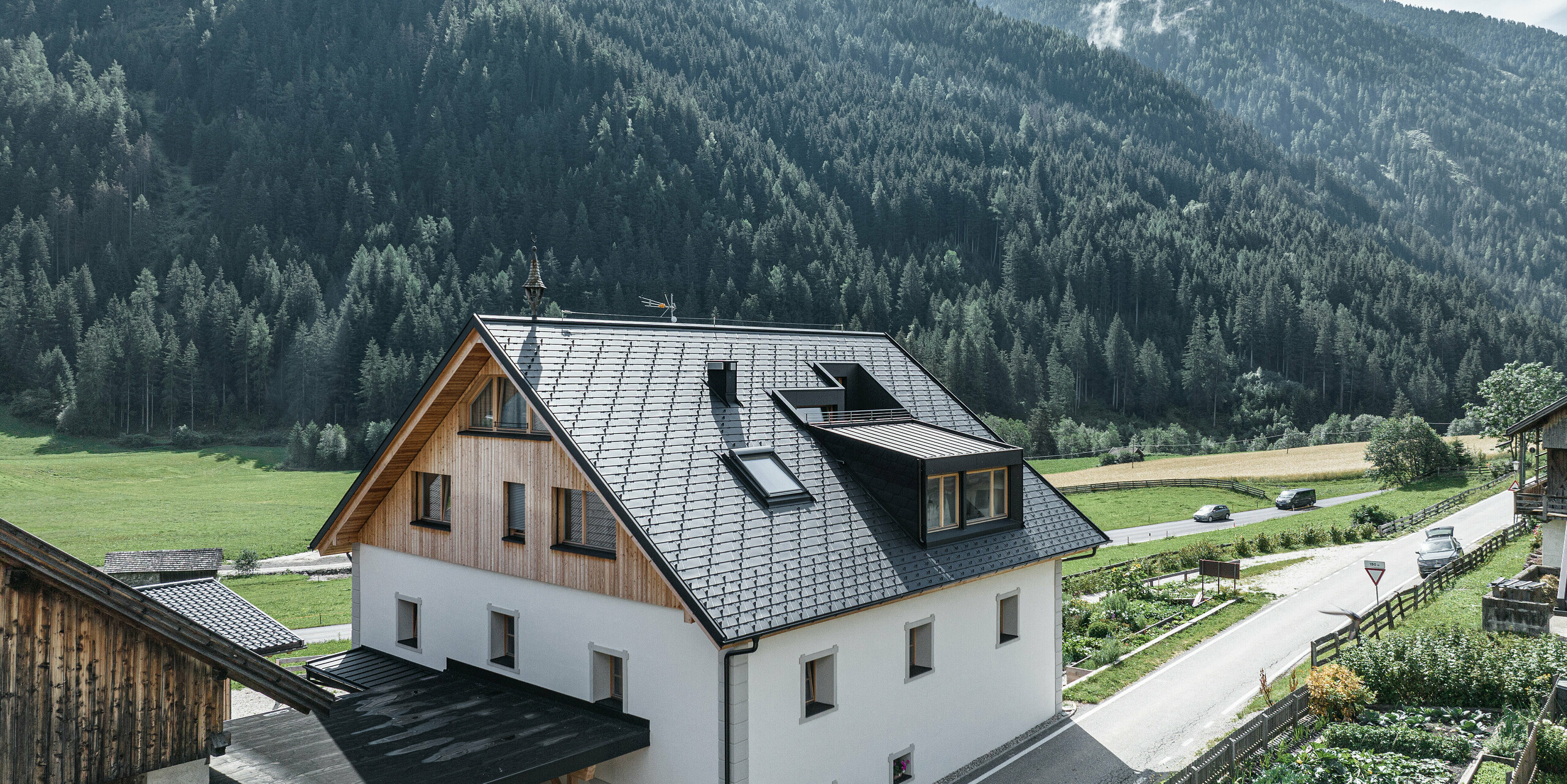 Perspektívny pohľad na Ausserwegerhof v Antholzi v Južnom Tirolsku s modernou strechou PREFA zo šindľov DS.19 antracitovej farby. Biela fasáda budovy vyčnieva z hustého ihličnatého lesa a tichej pouličnej scény. Štíty budovy sú obložené drevom a tvoria harmonický prechod k hliníkovej streche. Zo sedlovej strechy na oboch stranách vyčnieva vikier. Pod vikiermi sú priestranné okná, ktoré poskytujú dobrý výhľad. 
