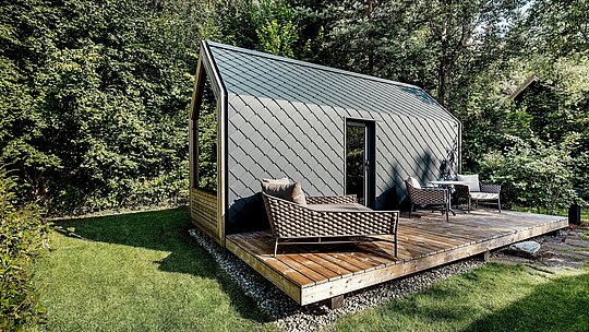 Minimalistický domček typu „tiny house“ v areáli Secret Koi Garden pri jazere Natterer See stojí na priestrannej drevenej terase, obklopený hustou lesnou vegetáciou. Strecha a fasáda sú vyhotovené z malých hliníkových strešných a fasádnych šablón PREFA v odtieni P.10 antracit, ktoré svojou jednotnou štruktúrou dotvárajú moderný vzhľad. Odolný a poveternostne odolný hliníkový plášť poskytuje spoľahlivú ochranu a vytvára silný kontrast s drevom, zeleňou a otvoreným vonkajším obytným priestorom glampingového rezortu.