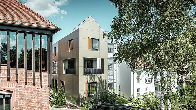 Na obrázku je moderná radnica v Alzenau, ktorej fasáda je vyrobená z hliníkových kompozitných dosiek PREFA v bronzovej farbe. Špicatá strecha a jasne štruktúrované okná s drevenými žalúziami dodávajú budove elegantný a moderný vzhľad. V popredí je vidieť zelená plocha s dláždenou cestičkou a stromami, ktorá harmonicky dopĺňa mestský dizajn budovy.