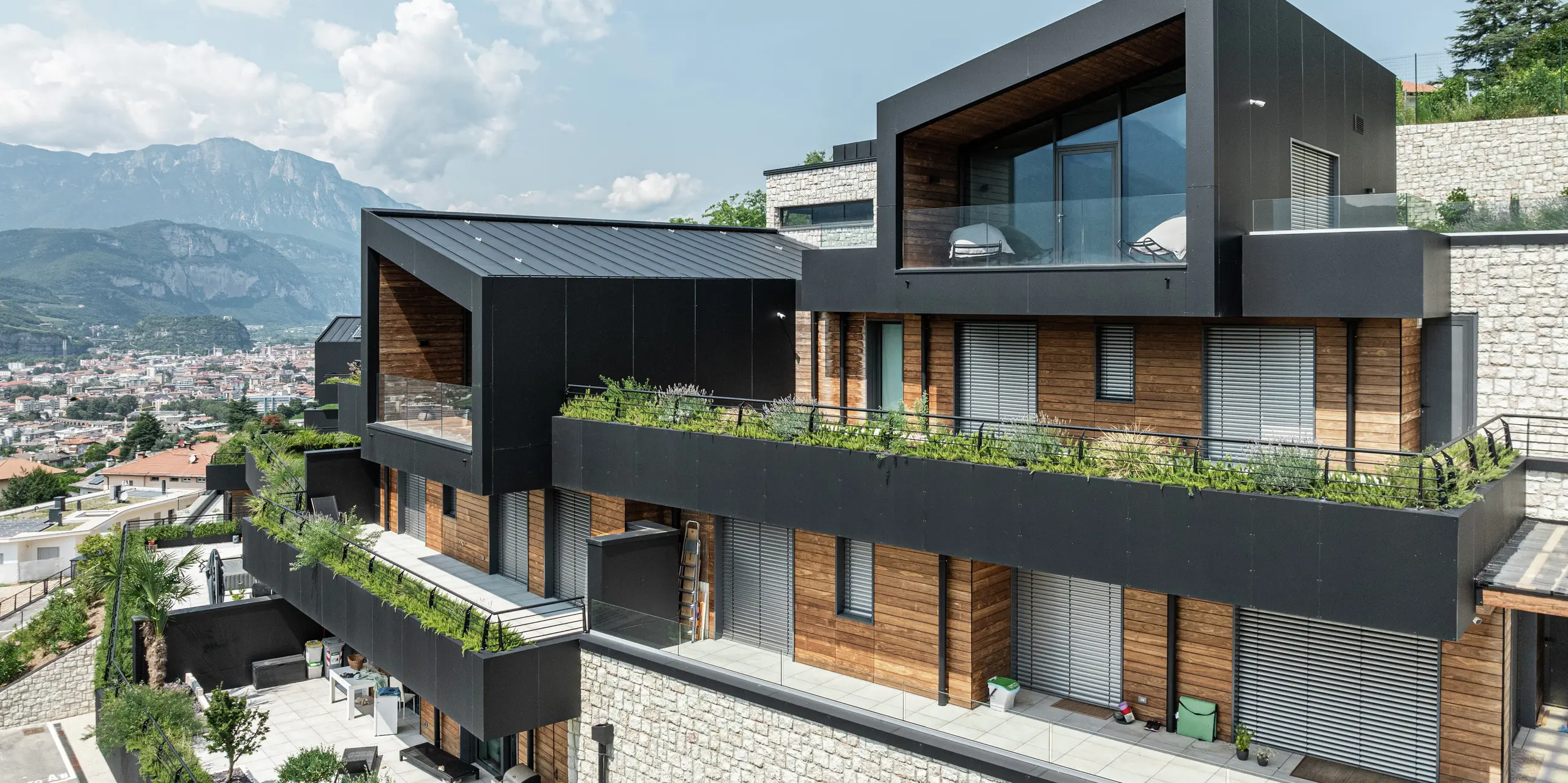 Moderná architektúra s charakterom: Elegantná fasáda z hliníkových kompozitných dosiek PREFA v čiernej farbe P.10 dokonale ladí s teplými drevenými prvkami a podstavcom z prírodného kameňa. Strecha PREFALZ v čiernej farbe P.10 ponúka nielen estetickú jednotnosť, ale aj najvyššiu odolnosť voči poveternostným vplyvom. Veľkorysé sklenené plochy a zelené terasy vytvárajú dokonalé prepojenie medzi vnútorným a vonkajším priestorom, zatiaľ čo poloha na svahu umožňuje impozantný výhľad na Trident. Trvanlivý, nenáročný na údržbu a štýlový – koncept bývania s budúcnosťou.