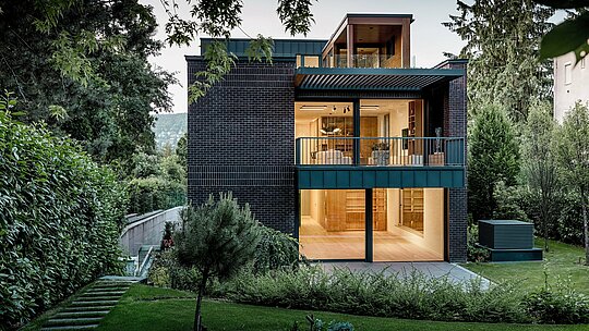 Zadná strana modernej viacgeneračnej vily v Budapešti je zasadená do bohato zelenej záhradnej krajiny. Veľké presklené plochy otvárajú obytný priestor smerom von a umožňujú, aby teplé vnútorné svetlo dopadalo na štruktúrovanú tehlovú fasádu. Čisté línie budovy zvýrazňuje hliník PREFALZ v odtieni P.10 antracit, ktorý ako obklad balkónov a fasádny prvok vytvára trvácne akcenty. Kombinácia tehly, hliníka a bohatej zelene podčiarkuje nadčasový, kvalitný a architektonicky náročný charakter domu.