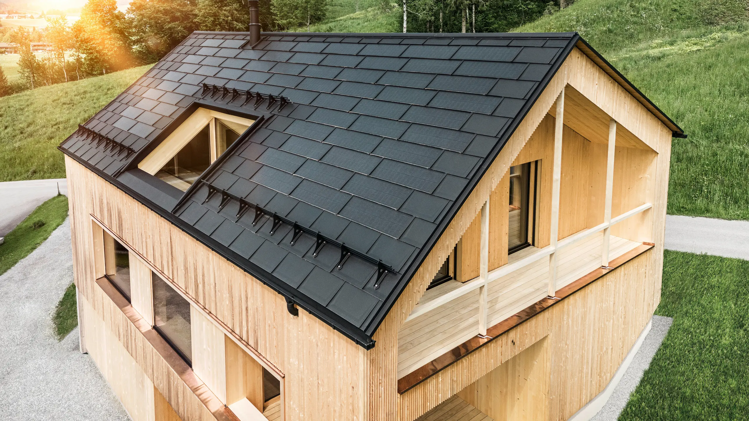 [Translate to Slovenčina:] Blick auf ein modernes, nachhaltiges Holzhaus in Egg mit einem PREFA Solardach, das durch seine dunkle, strukturierte sowie elegante Erscheinung auffällt. Die Solarmodule sind strategisch zwischen den klassischen Dachplatten R.16 platziert, um eine optimale Sonneneinstrahlung zu nutzen und sich dabei ästhetisch in das Gesamtdesign einzufügen. Das Haus steht im Einklang mit seiner grünen Umgebung, während die warme Abendsonne die natürliche Holztextur der Fassade unterstreicht und die solide Bauweise betont. Ein harmonisches Beispiel für umweltbewusstes Bauen und Energieeffizienz.